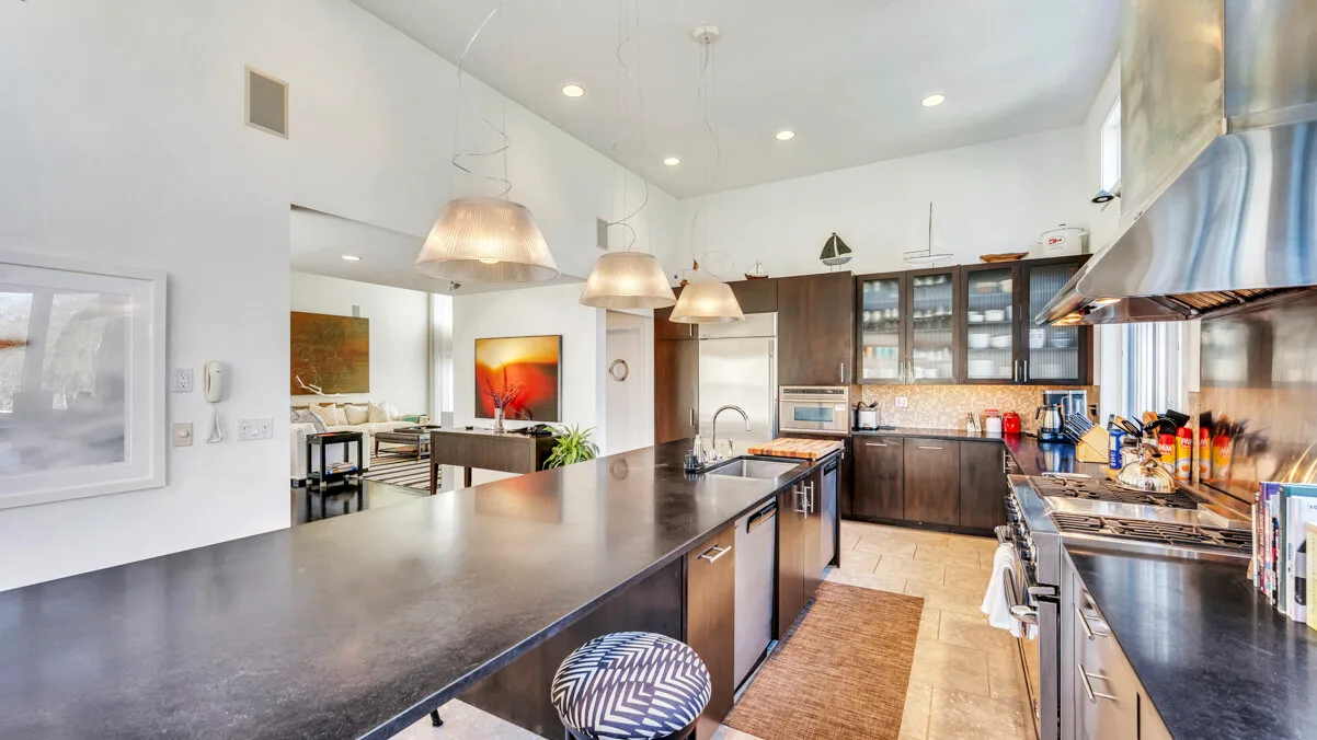 A bright kitchen with a long island, hanging lights, and dark cabinetry.