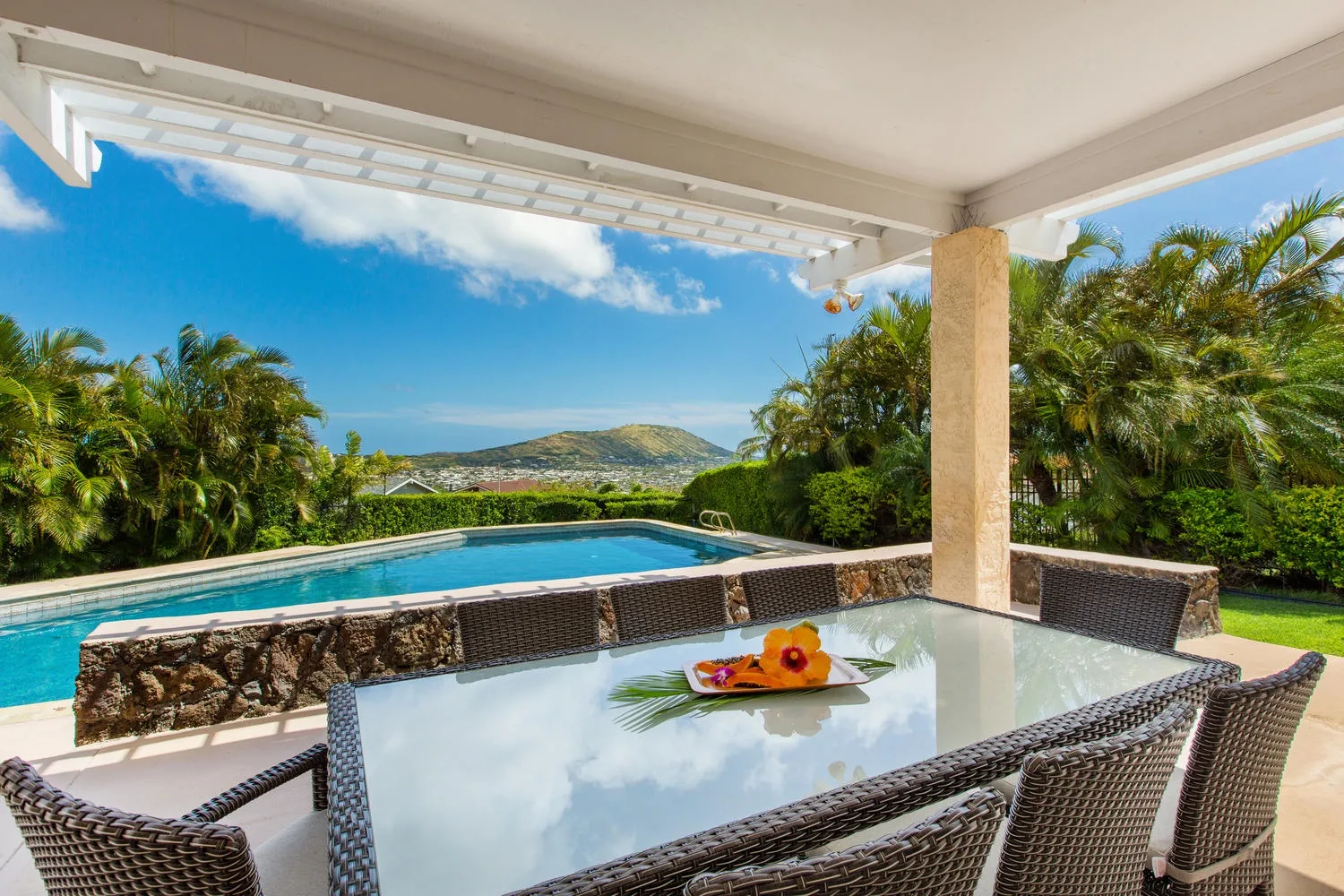 Un comedor exterior cubierto da a una piscina y una ladera verde.