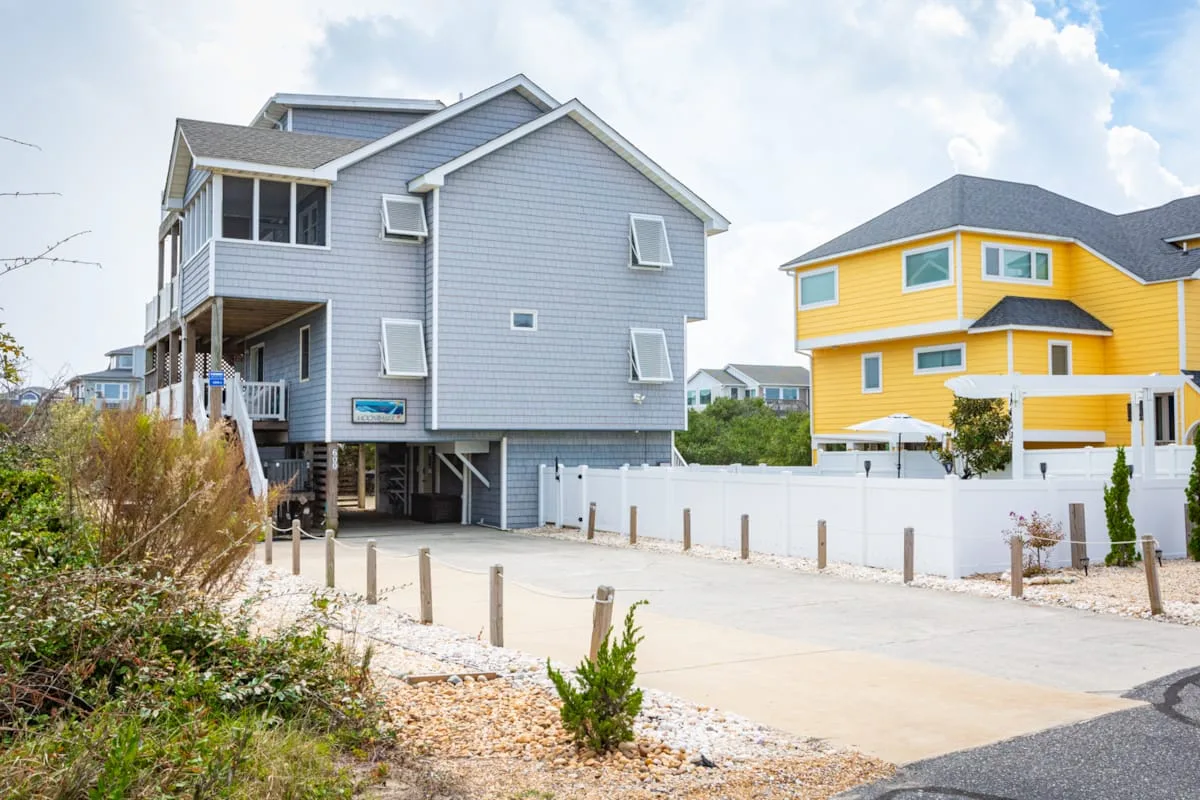 A gray coastal-style house stands beside a bright yellow house along a paved lane.