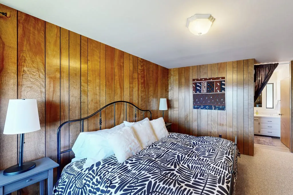 Dormitorio con una cama cubierta con ropa de cama estampada y paredes revestidas de madera.