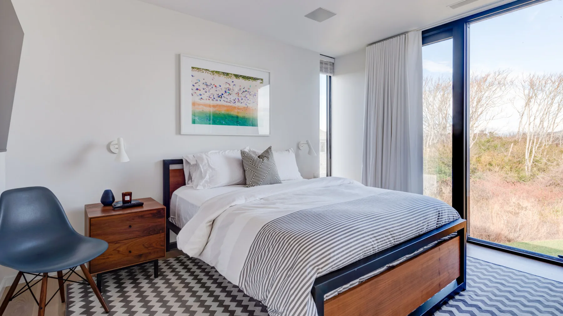 A bedroom with a neatly made bed beside large windows and a bedside table.