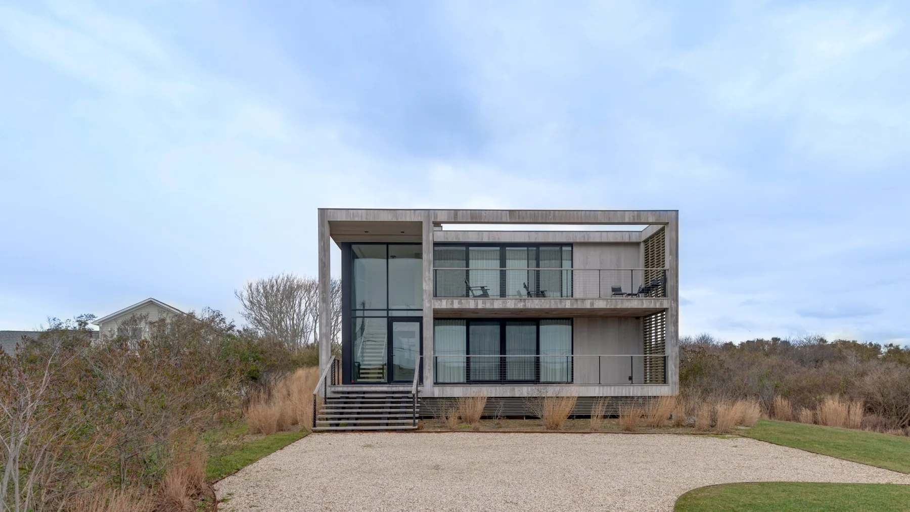 A modern glass-fronted house stands beside a gravel driveway and open lawn.