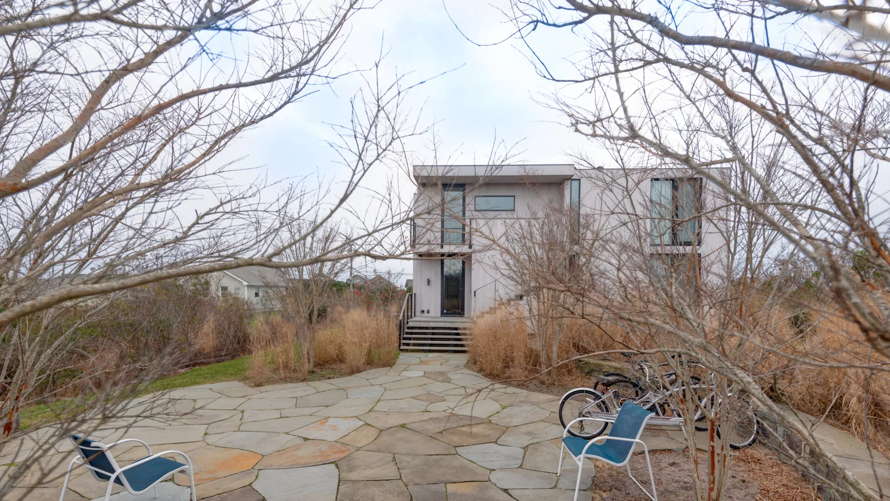 A modern house stands beyond a stone-paved path, framed by bare trees.