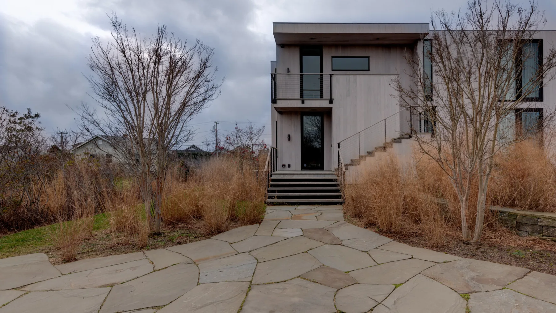 A modern house stands beyond a stone-paved path, surrounded by dry grasses and bare trees.