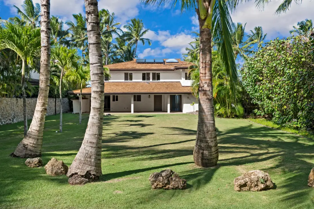 A two-story house stands behind a lawn shaded by palm trees.
