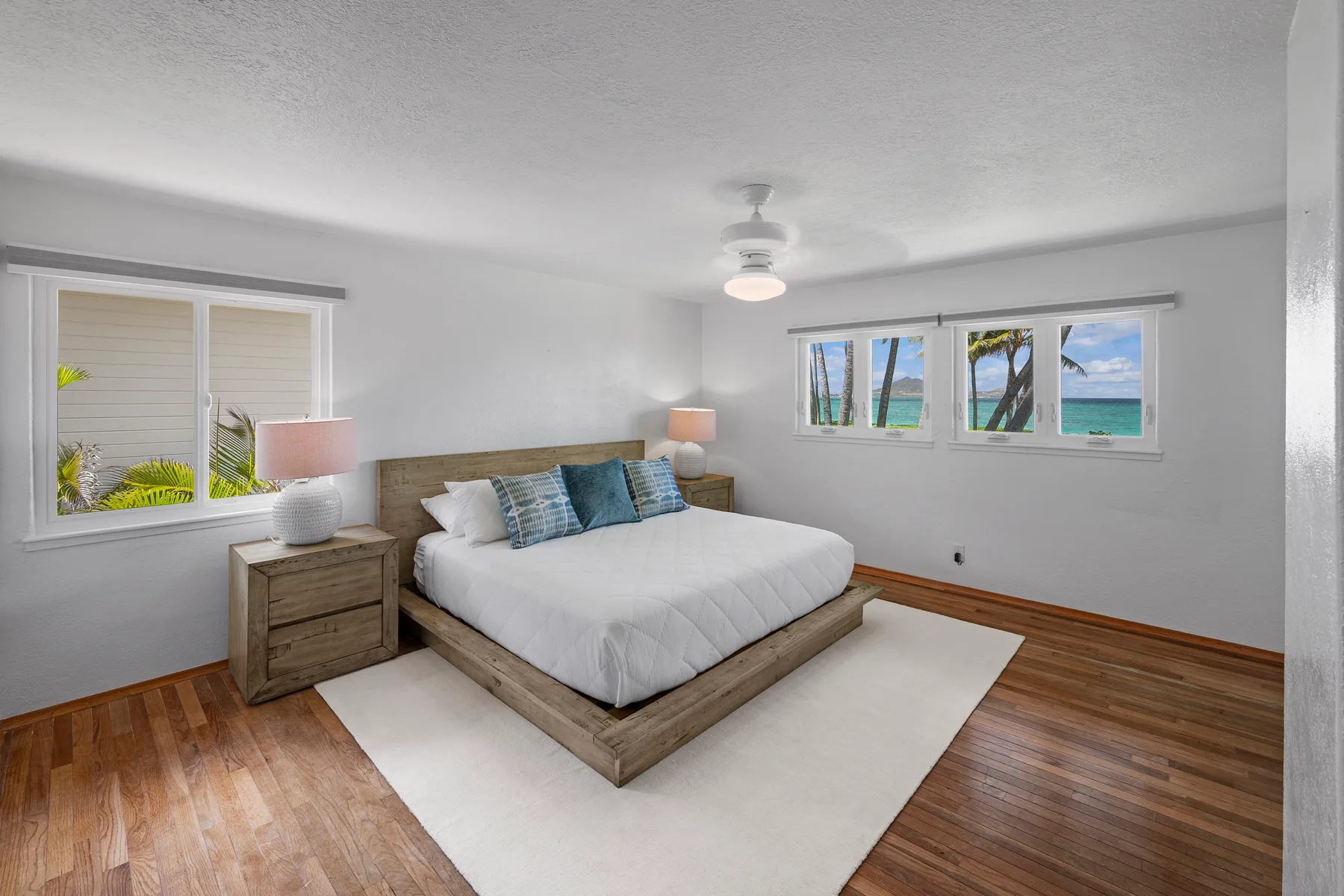 A bright bedroom has a bed with blue pillows and windows overlooking the water.