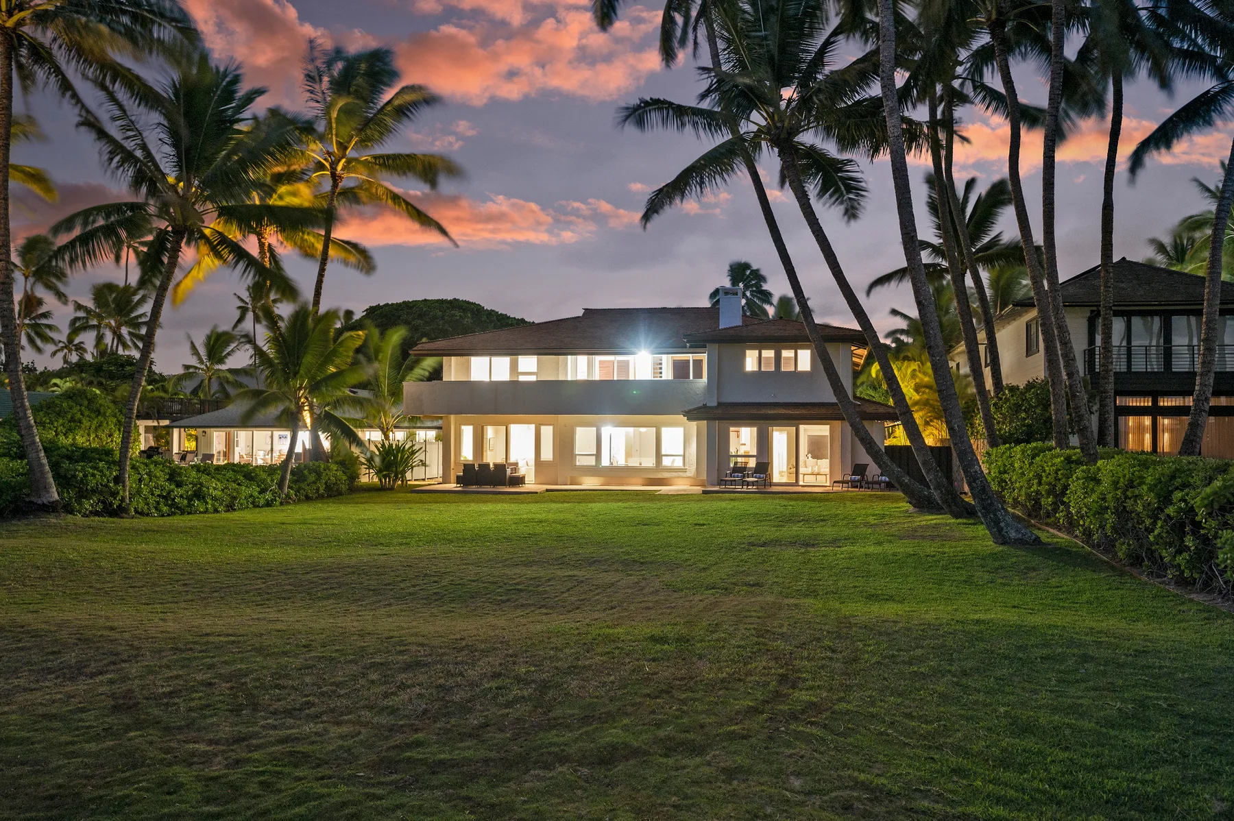 Una casa grande con ventanas iluminadas se alza detrás de un césped y palmeras al atardecer.