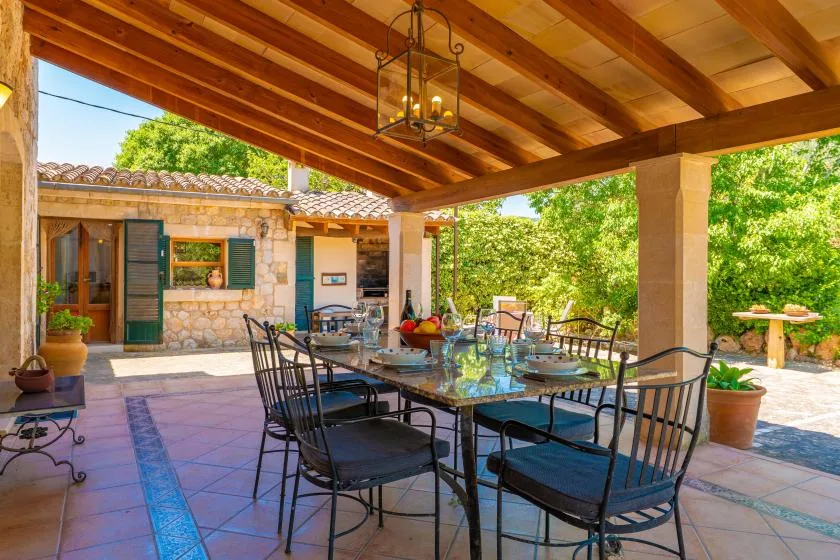 A covered outdoor dining area has a long table and seating beneath exposed wooden beams.