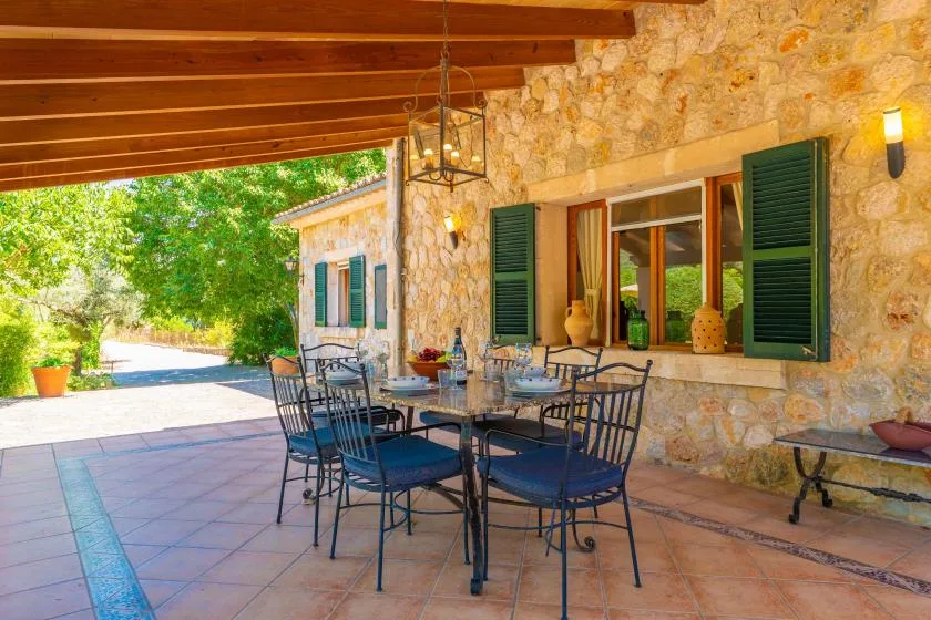 A shaded stone patio with a dining table and seating beside green-shuttered windows.