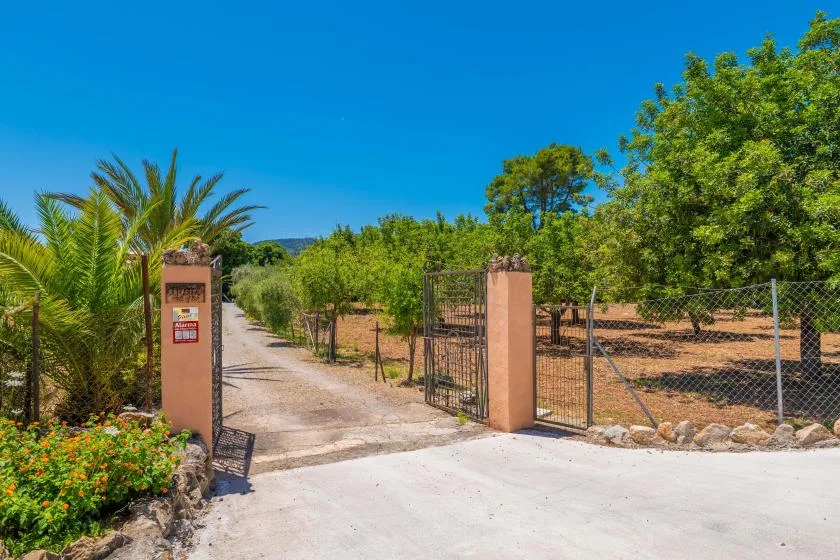 A gated rural entrance opens onto a dirt driveway bordered by trees.