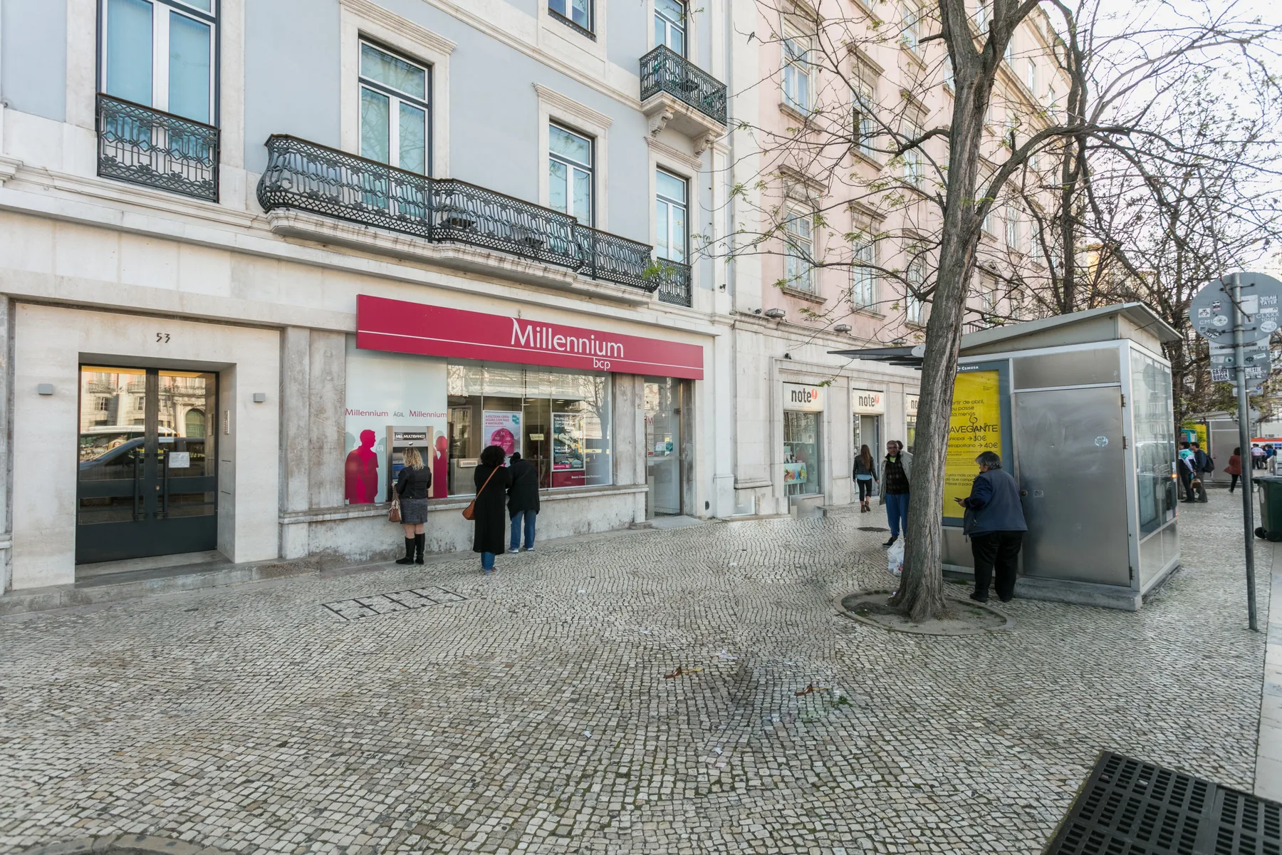 A cobblestone city street with a storefront, pedestrians, and a glass-sided shelter.