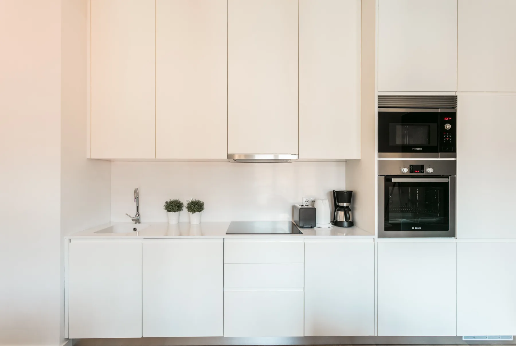 A modern white kitchen with built-in appliances and small potted plants on the counter.