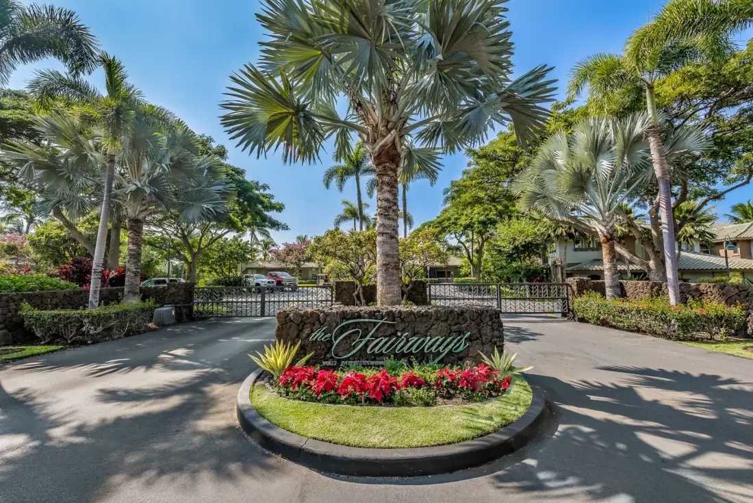 Una entrada ajardinada con una palmera central, macizos de flores y una entrada con portón.