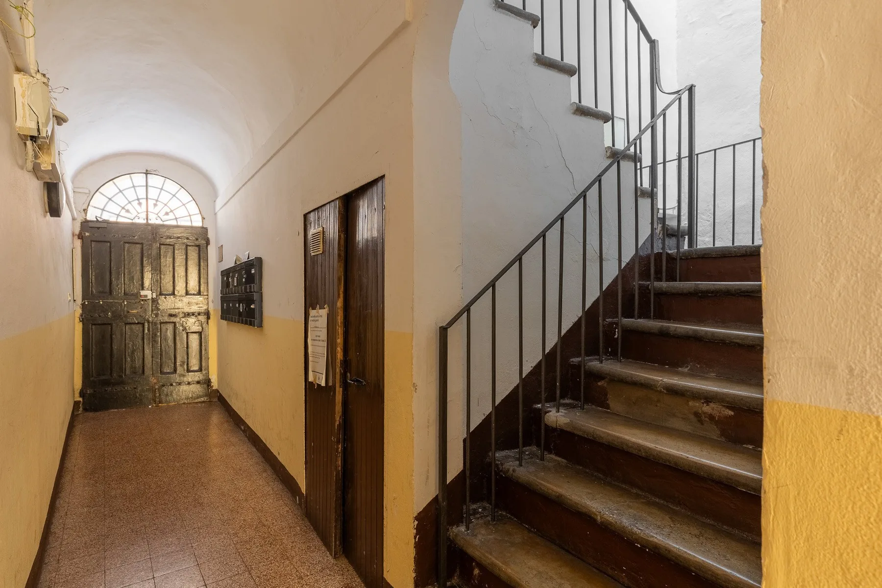 An interior hallway leads to a dark wooden door, beside a staircase with metal railings.
