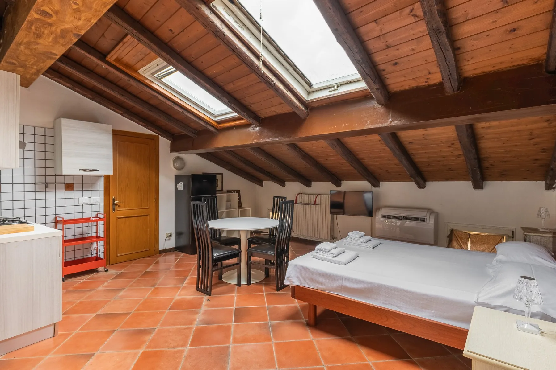 An attic bedroom with a bed, dining table, and skylights beneath a sloped wooden ceiling.