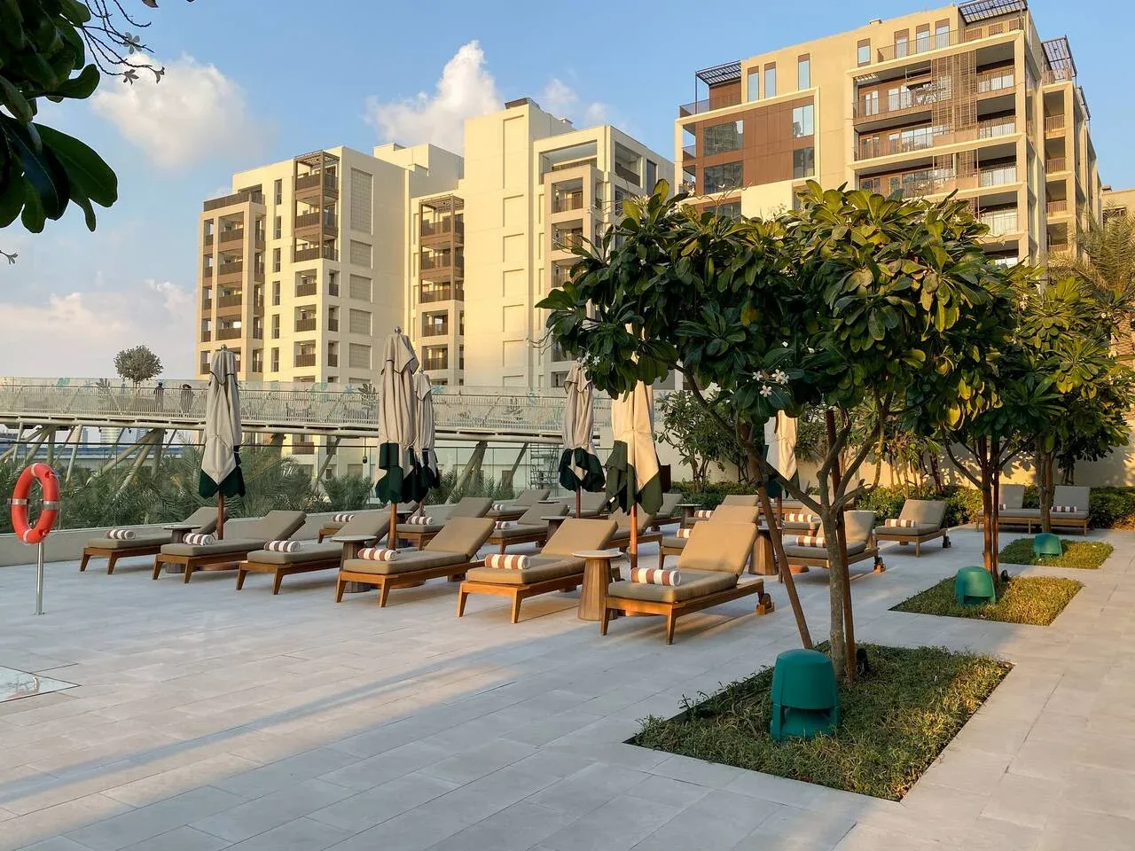 Una zona exterior junto a la piscina, con tumbonas y árboles frente a edificios de apartamentos.