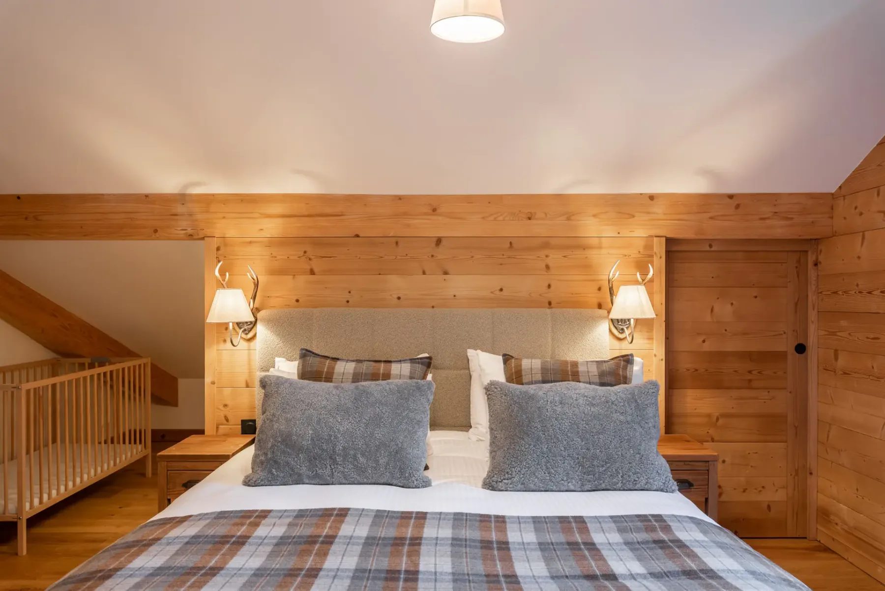 A cozy ensuite bedroom with wood-paneled walls, a double bed with grey and plaid bedding, bedside tables, a mirror, and a balcony door letting in natural light.