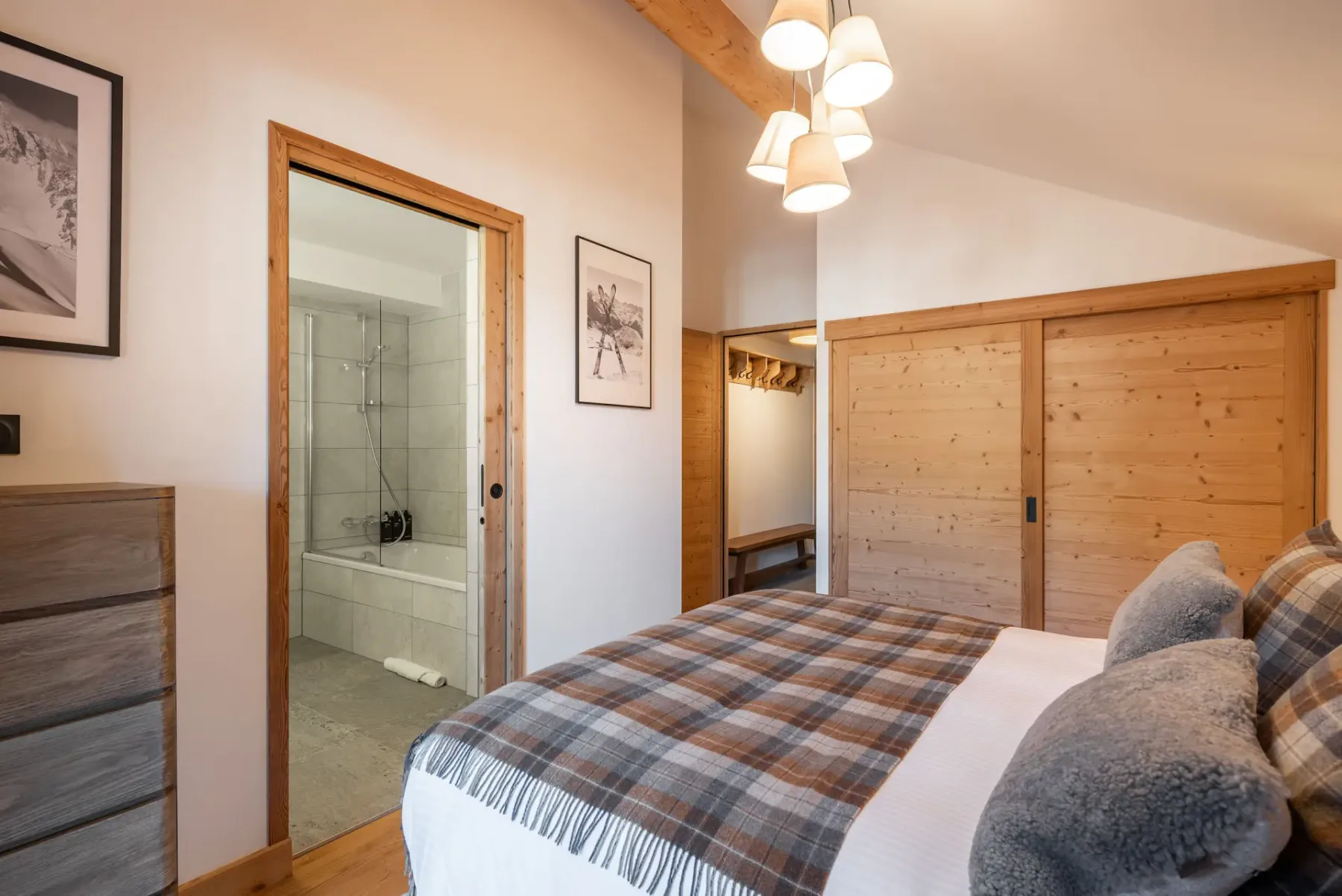 A cozy ensuite bedroom with wood-paneled walls, a double bed with grey and plaid bedding, bedside tables, a mirror, and a balcony door letting in natural light.