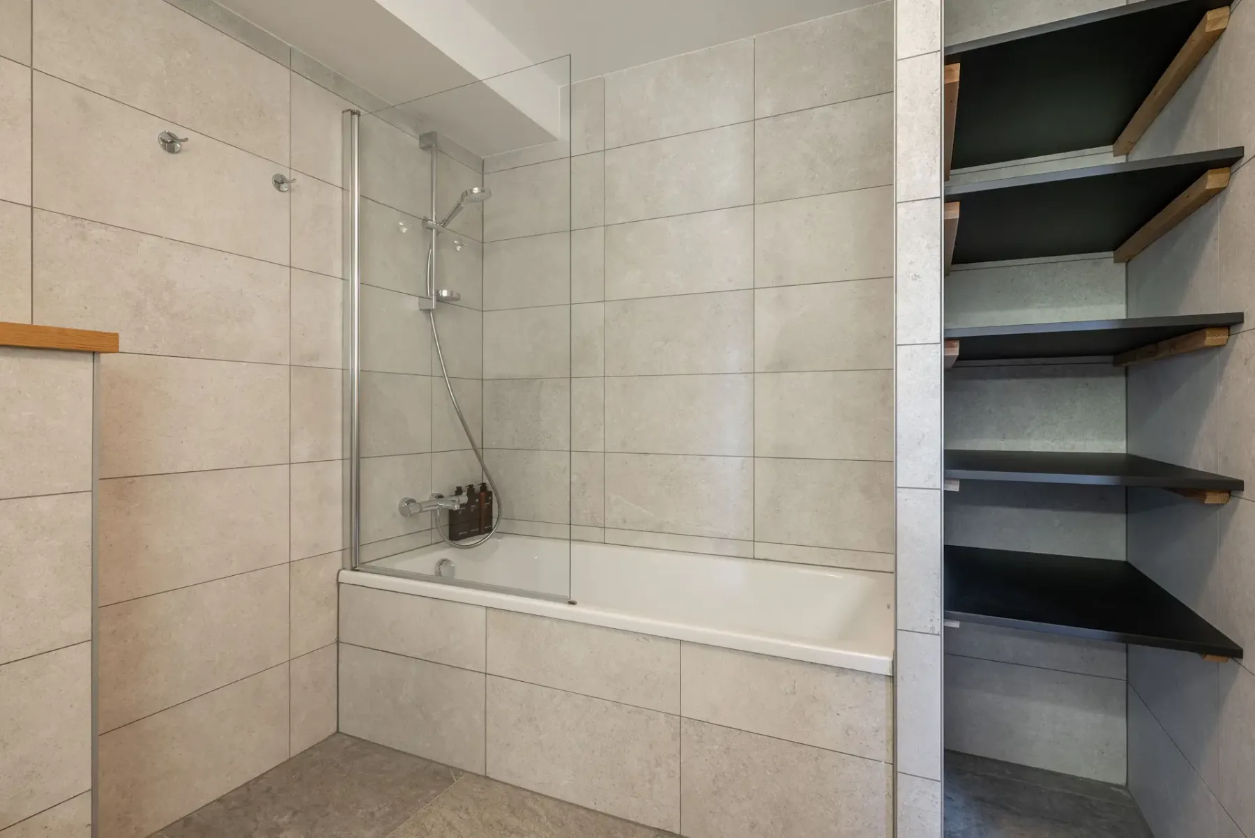 Modern bathroom with gray tile walls, wall-mounted toilet, glass-enclosed bathtub and shower, sink on a wooden vanity, and open shelving.