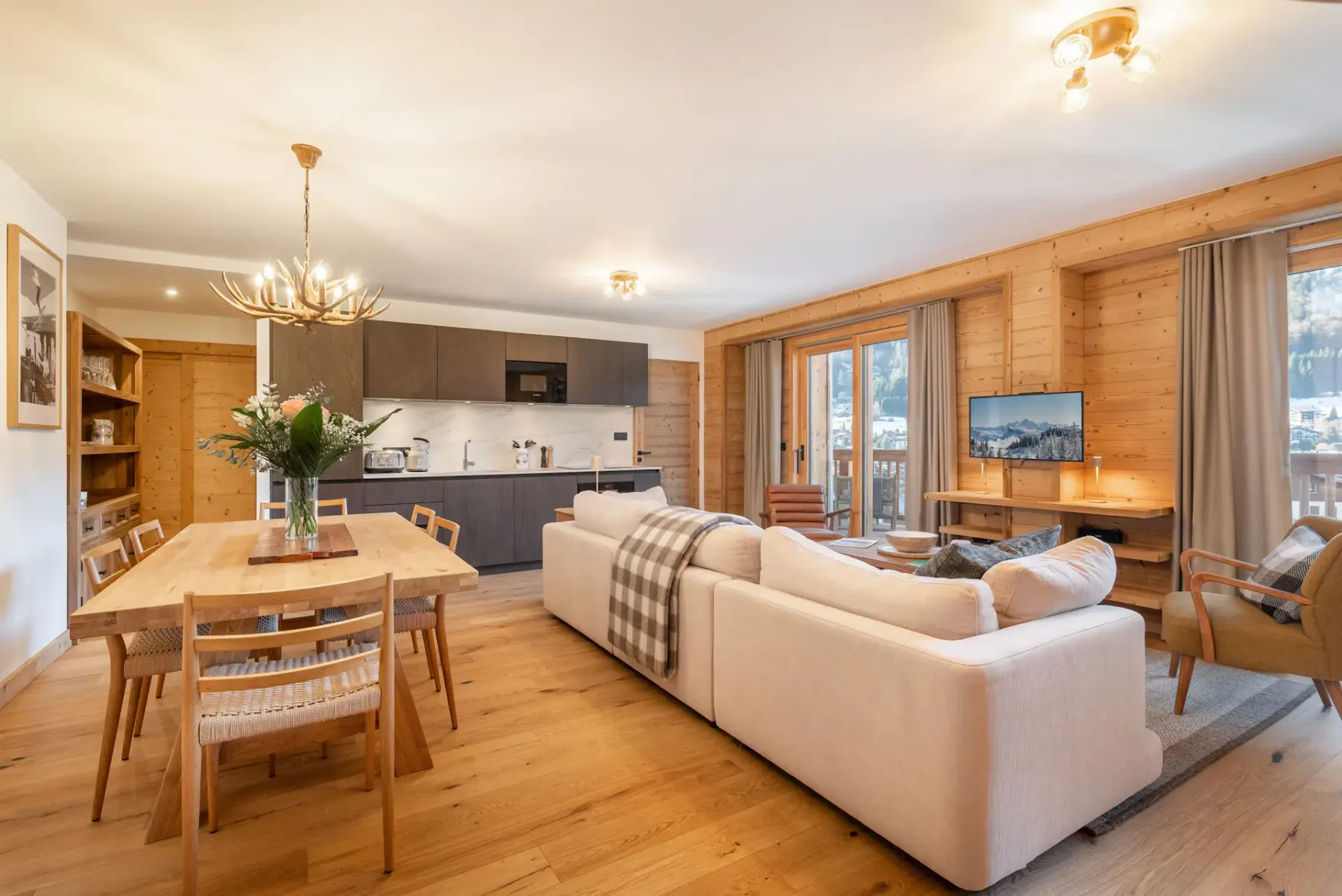 Open-plan living area with a beige sectional sofa, dining table, modern kitchen, wooden flooring and walls, and a staircase, with glass doors leading to a balcony and mountain view.