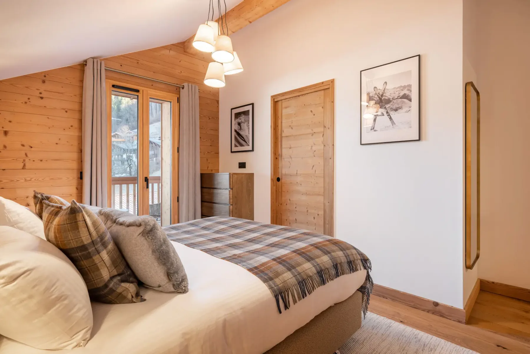 A cozy ensuite bedroom with wood-paneled walls, a double bed with grey and plaid bedding, bedside tables, a mirror, and a balcony door letting in natural light.