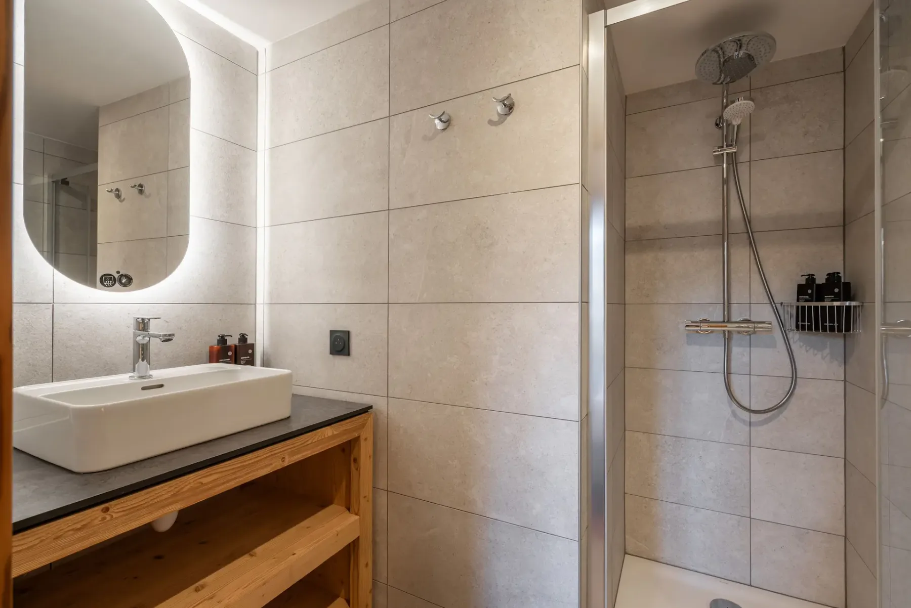 Modern bathroom with a wall-mounted sink, wooden open shelving, and a glass-enclosed shower with a rainfall showerhead and toiletries.