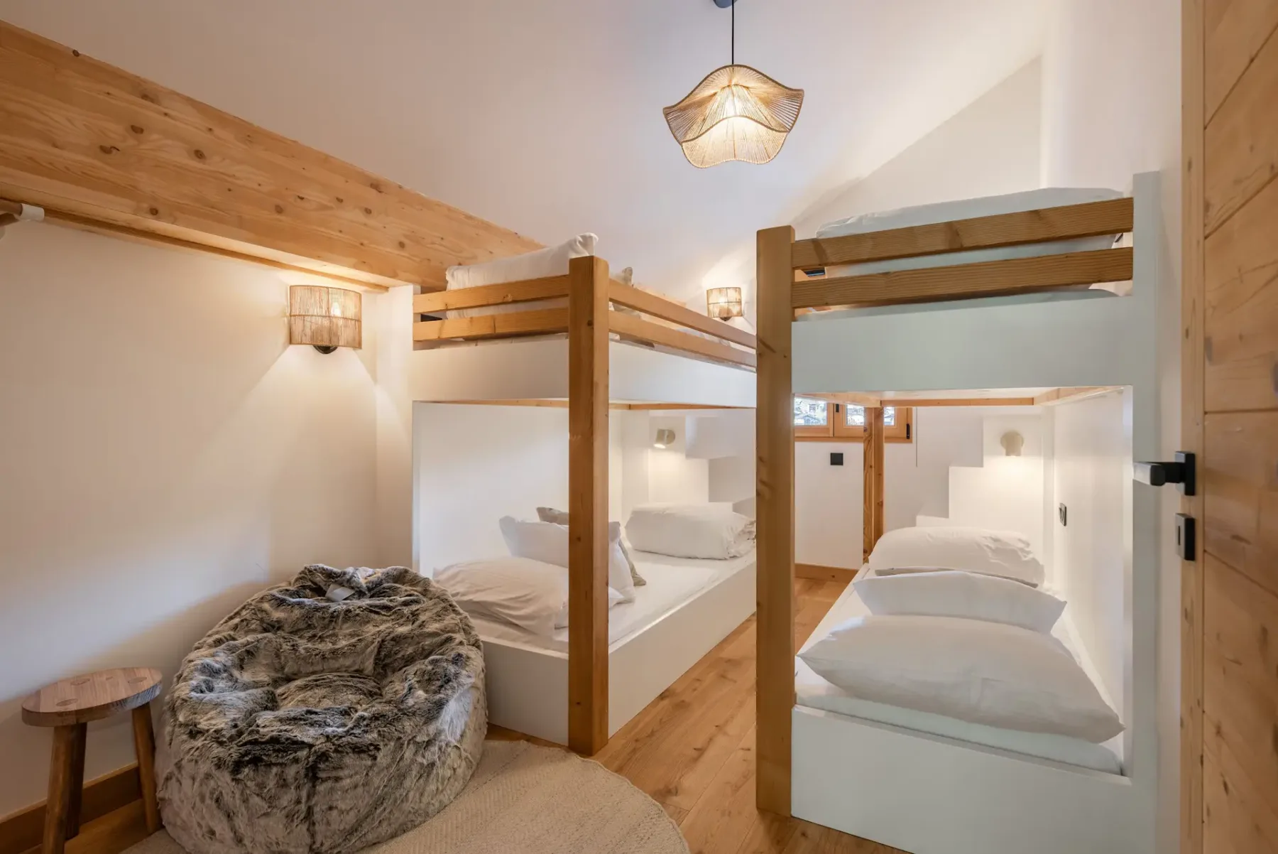 A cozy bedroom with two wooden bunk beds, white bedding, a round furry bean bag chair, a small wooden stool, and warm ambient lighting.