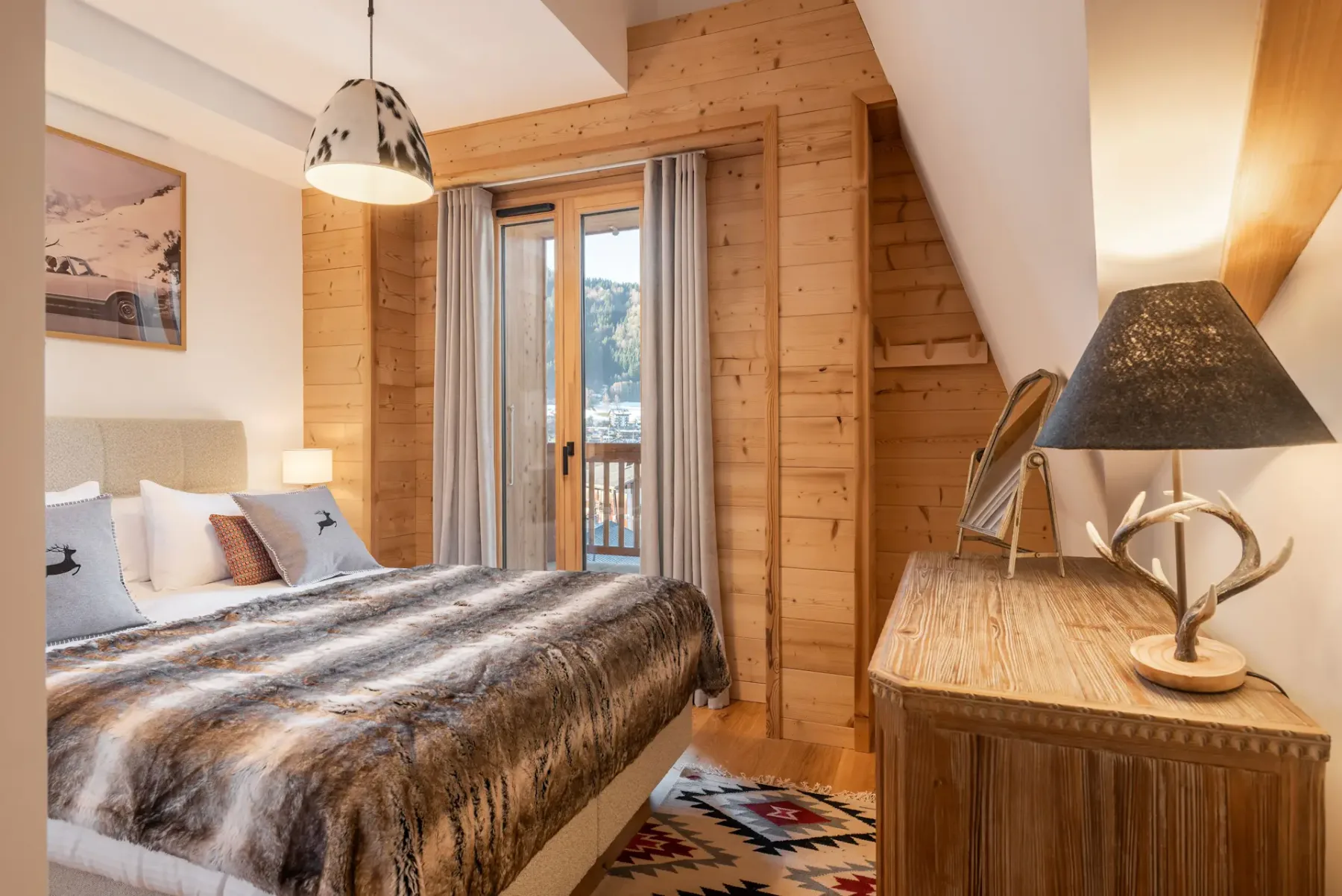 Cozy ensuite bedroom with wooden walls, a bed with fur blanket, a dresser with a lamp and mirror, and a window with mountain view.