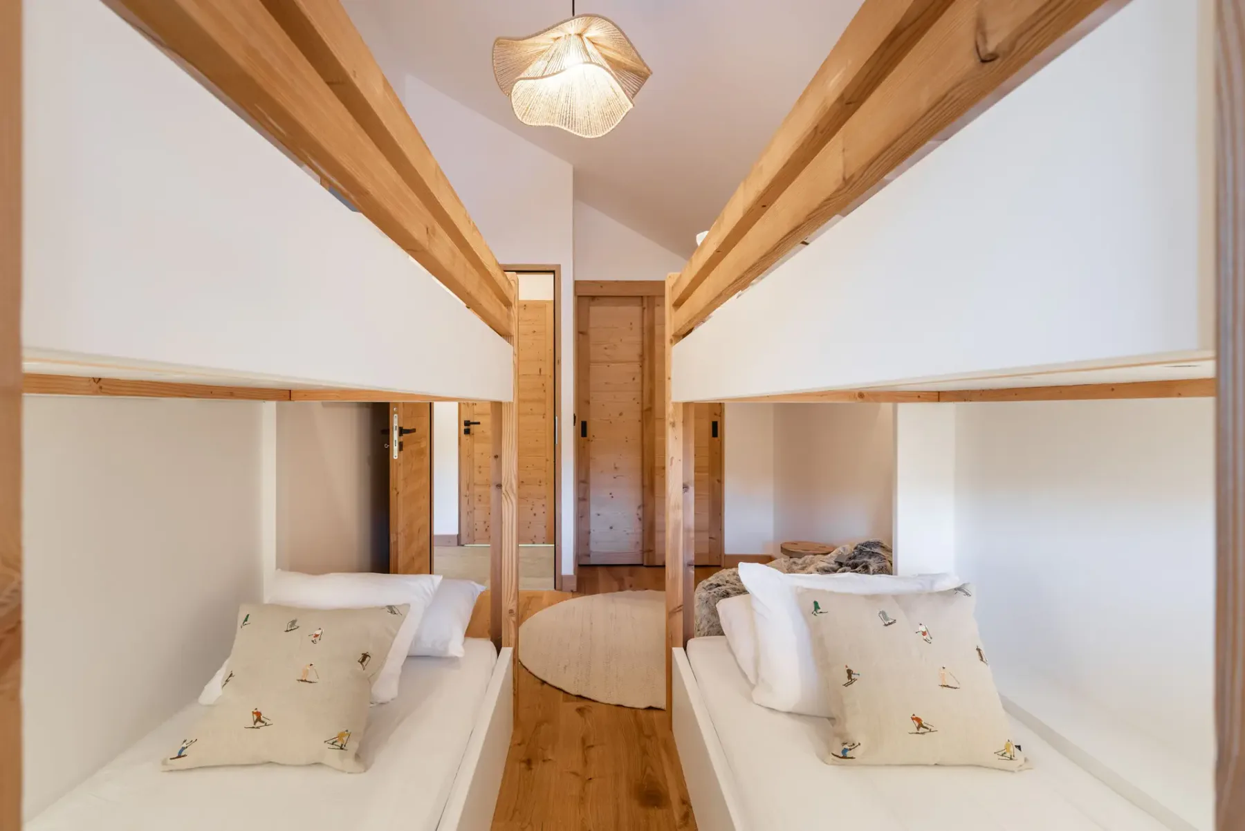 A cozy bedroom with two wooden bunk beds, white bedding, a round furry bean bag chair, a small wooden stool, and warm ambient lighting.