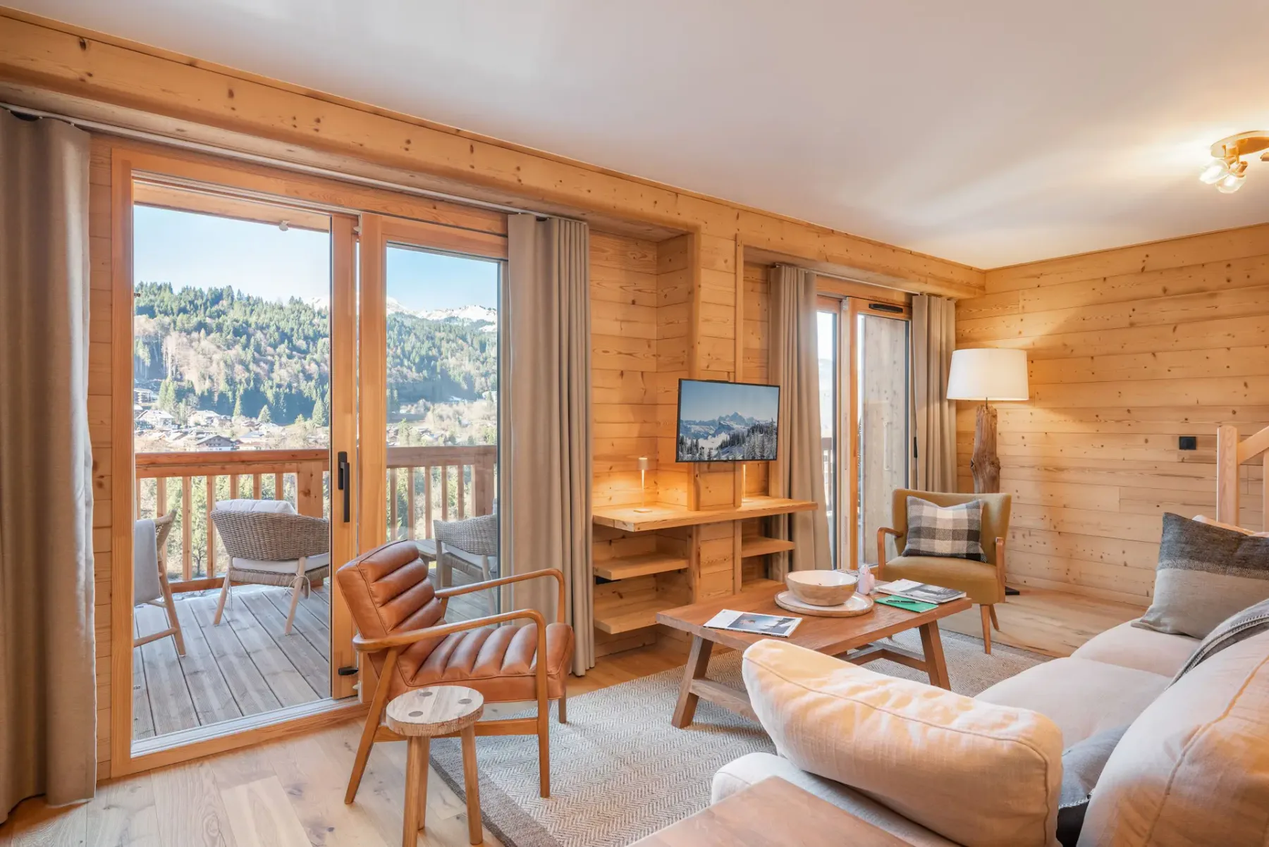 Open-plan living area with a beige sectional sofa, dining table, modern kitchen, wooden flooring and walls, and a staircase, with glass doors leading to a balcony and mountain view.