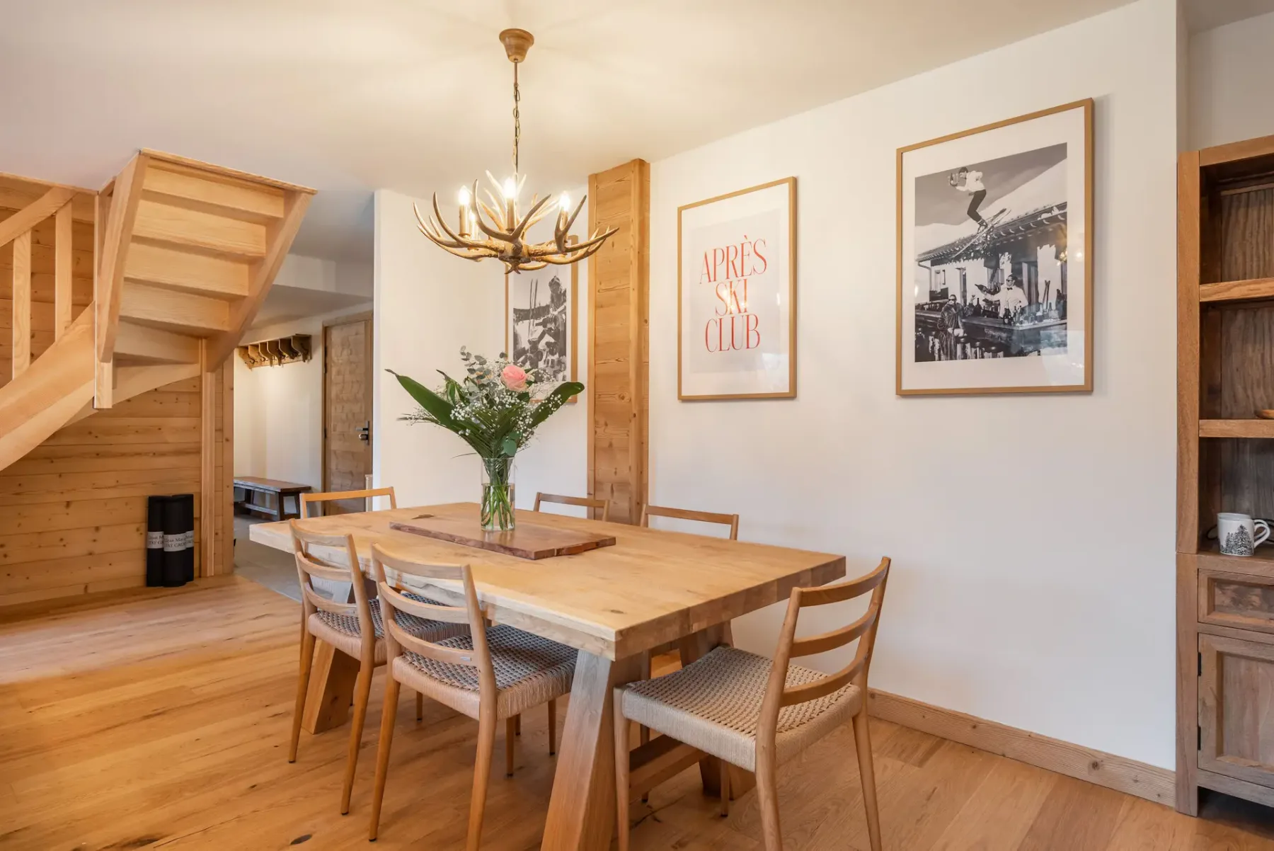 Modern dining area with a wooden table and six chairs, minimalist decor, flower centerpiece, wall art, and open kitchen with dark cabinets in the background.