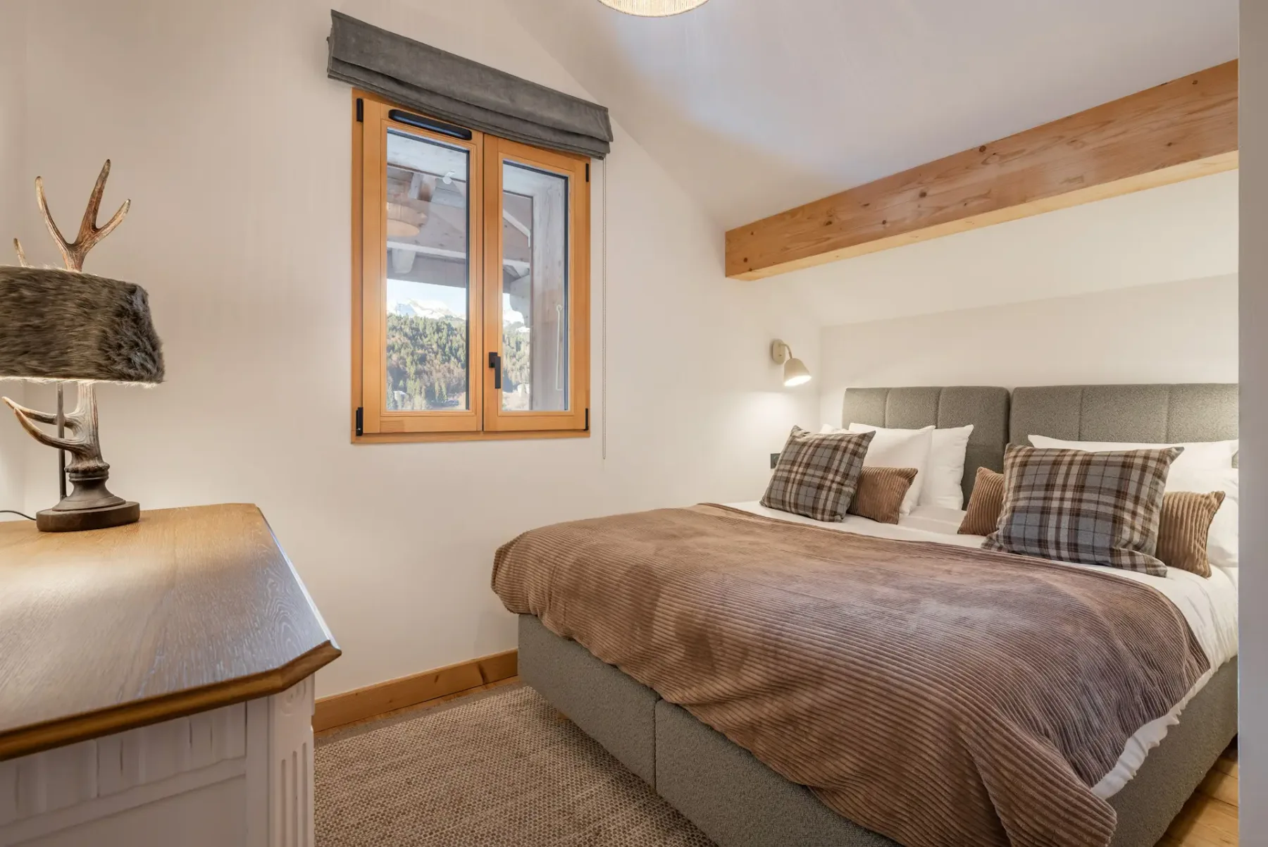 A cozy bedroom with a double bed, plaid pillows, a wooden beam ceiling, a window with a view, and a dresser with a lamp.