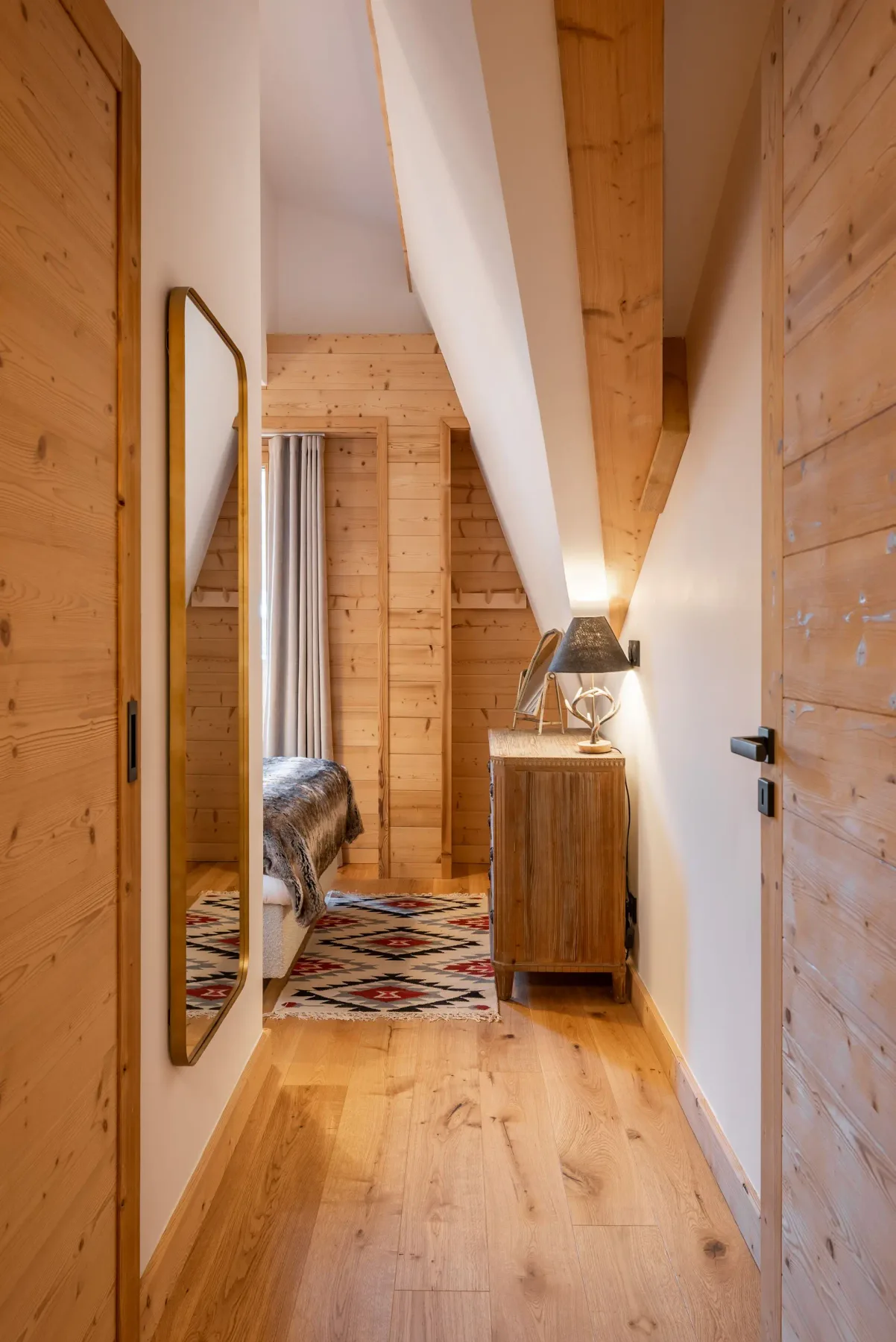 Cozy ensuite bedroom with wooden walls, a bed with fur blanket, a dresser with a lamp and mirror, and a window with mountain view.