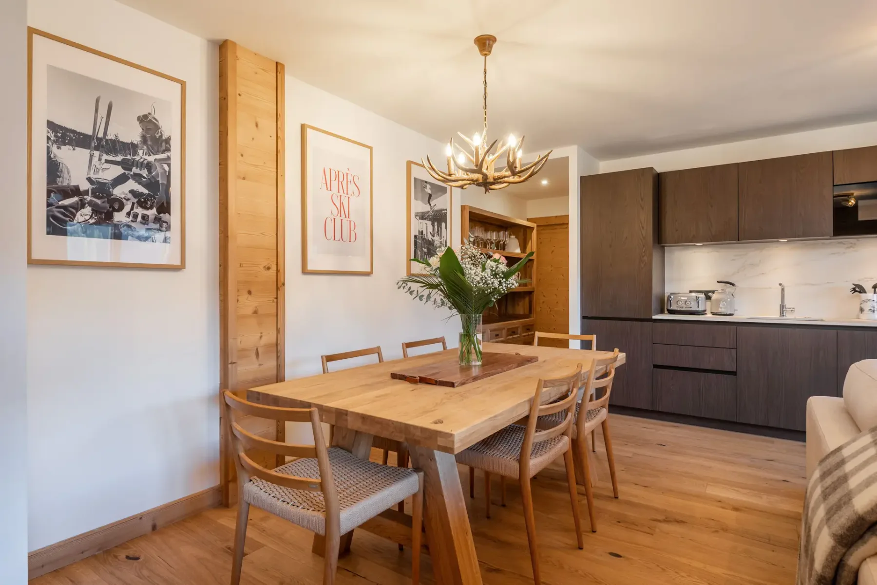 Modern dining area with a wooden table and six chairs, minimalist decor, flower centerpiece, wall art, and open kitchen with dark cabinets in the background.