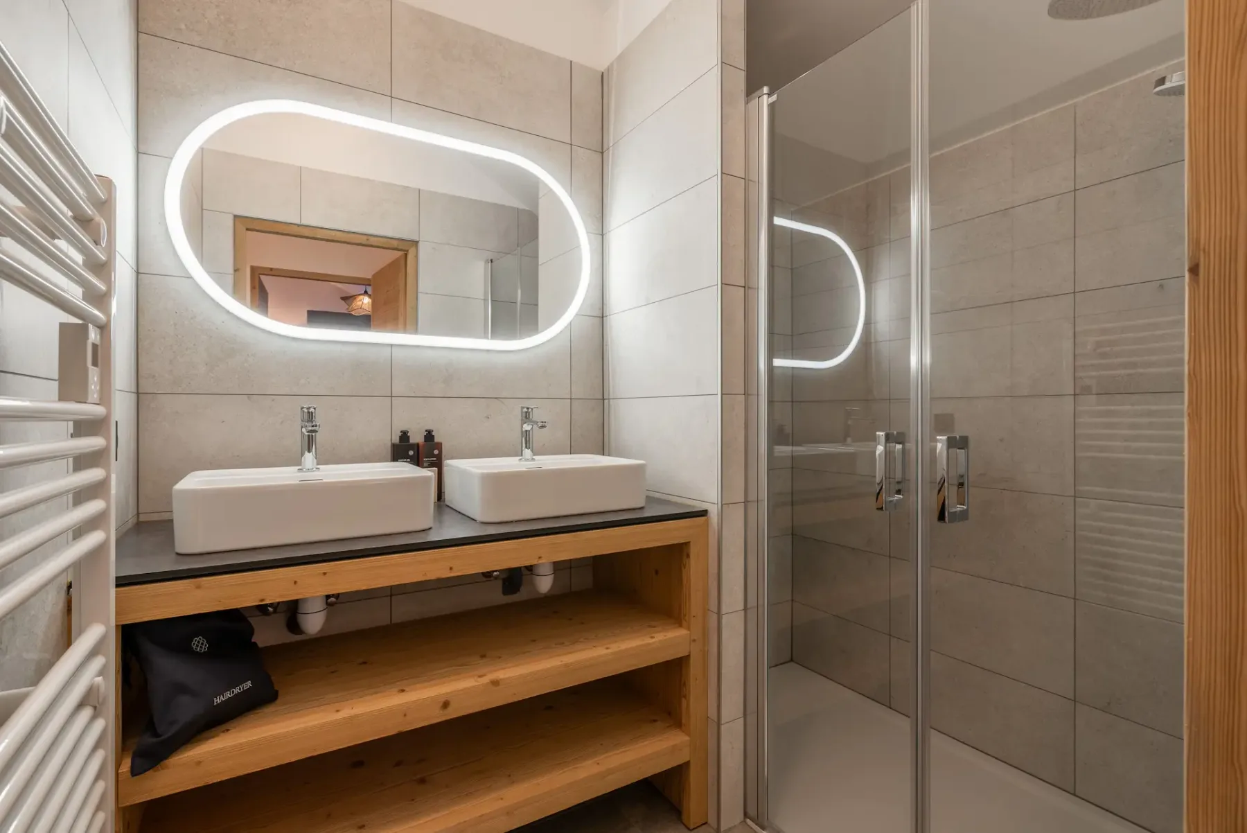 Modern bathroom with double sinks, a large illuminated mirror, wooden shelving, and a glass-enclosed walk-in shower.