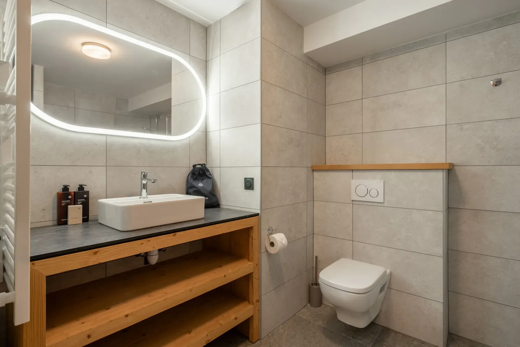 Modern bathroom with gray tile walls, wall-mounted toilet, glass-enclosed bathtub and shower, sink on a wooden vanity, and open shelving.