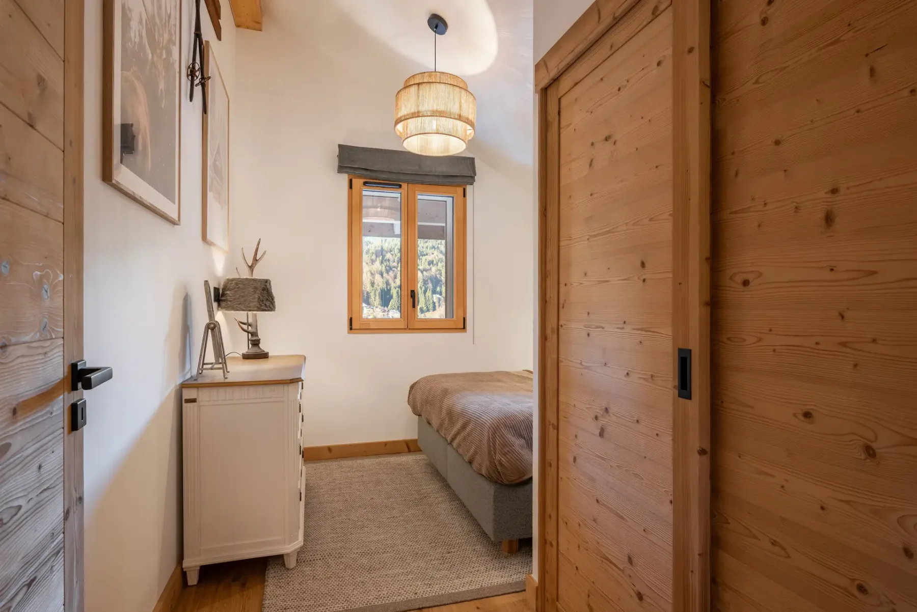 A cozy bedroom with a double bed, plaid pillows, a wooden beam ceiling, a window with a view, and a dresser with a lamp.