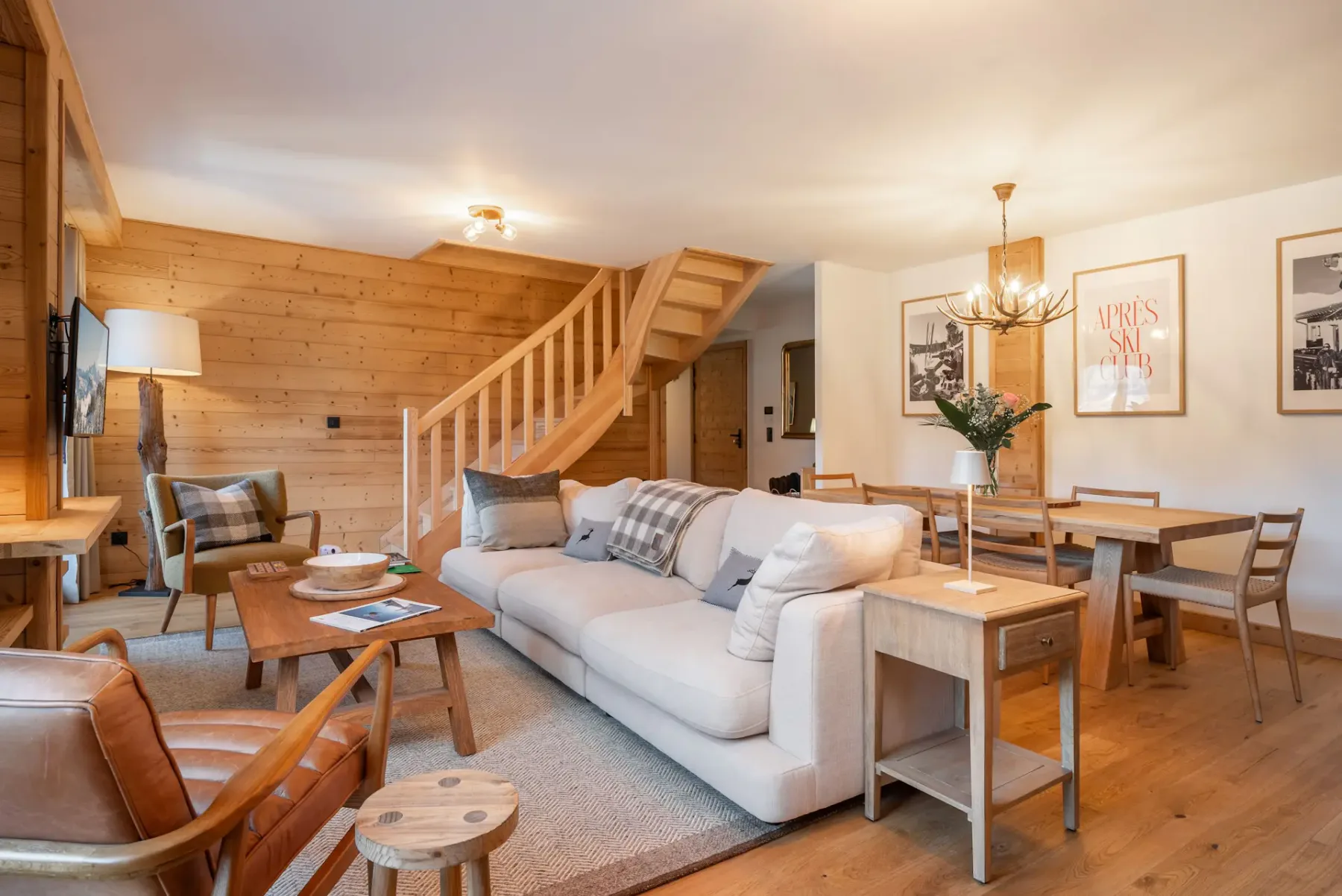 Open-plan living area with a beige sectional sofa, dining table, modern kitchen, wooden flooring and walls, and a staircase, with glass doors leading to a balcony and mountain view.