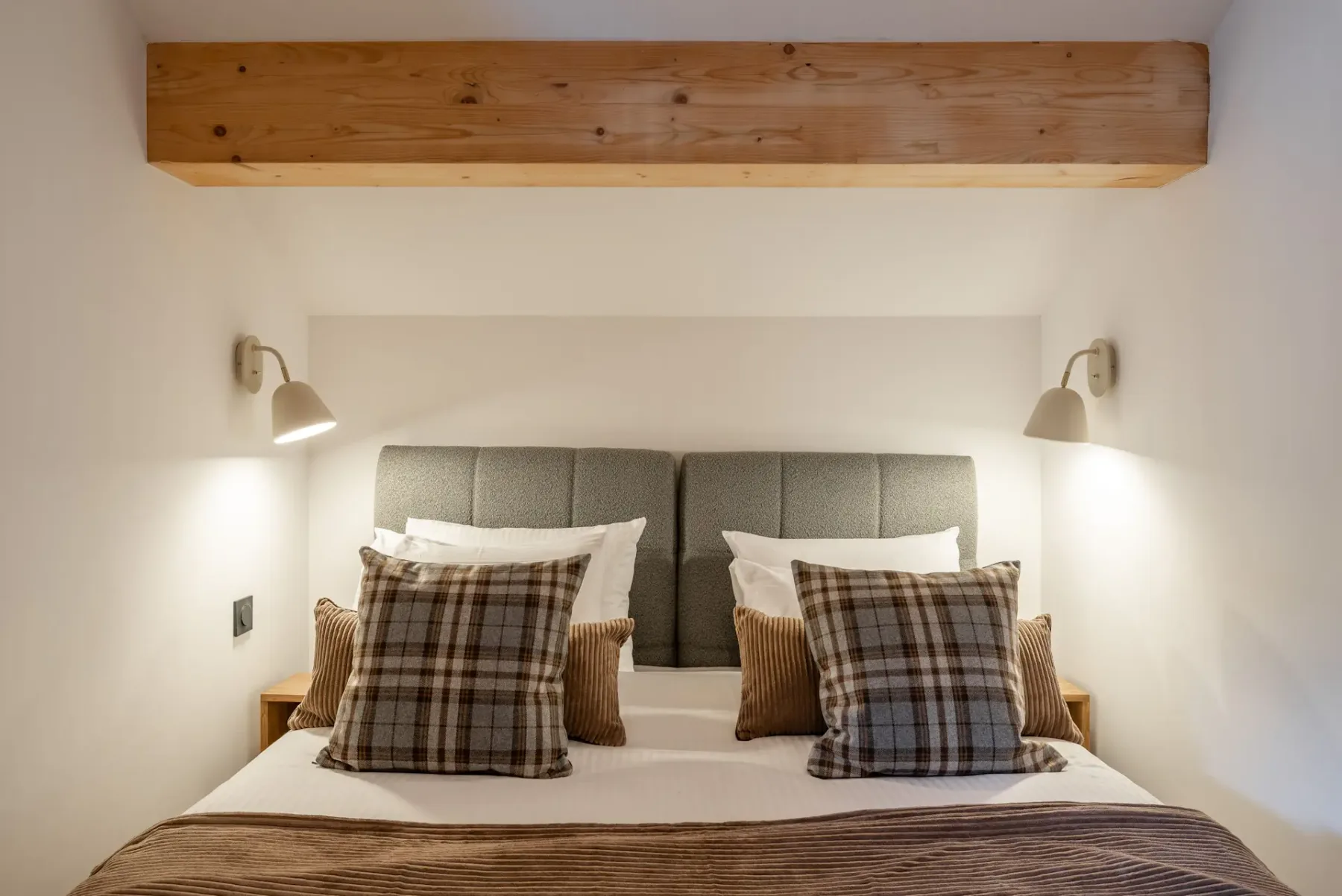 A cozy bedroom with a double bed, plaid pillows, a wooden beam ceiling, a window with a view, and a dresser with a lamp.