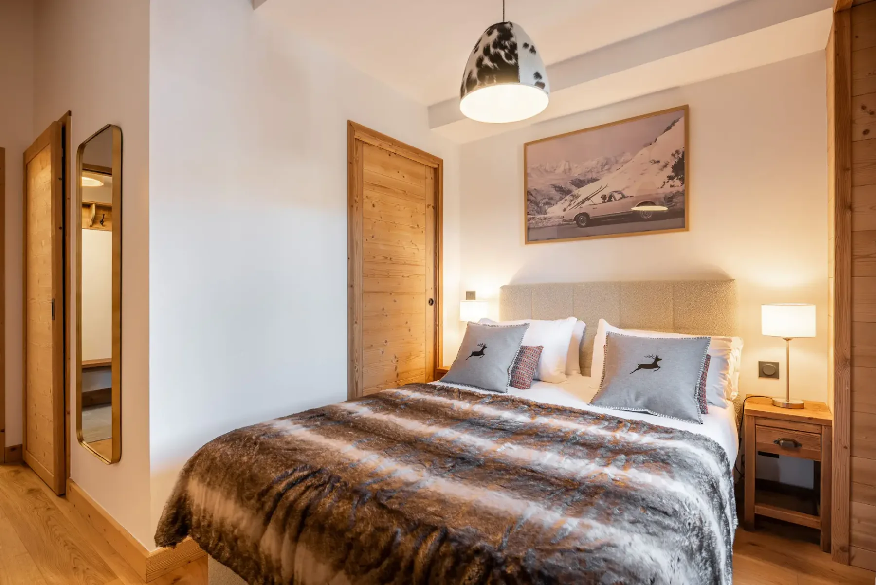 Cozy ensuite bedroom with wooden walls, a bed with fur blanket, a dresser with a lamp and mirror, and a window with mountain view.