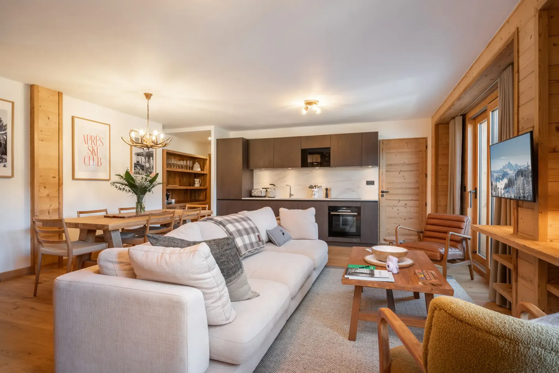 Open-plan living area with a beige sectional sofa, dining table, modern kitchen, wooden flooring and walls, and a staircase, with glass doors leading to a balcony and mountain view.