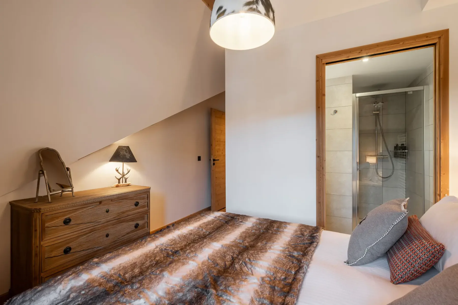 Cozy ensuite bedroom with wooden walls, a bed with fur blanket, a dresser with a lamp and mirror, and a window with mountain view.