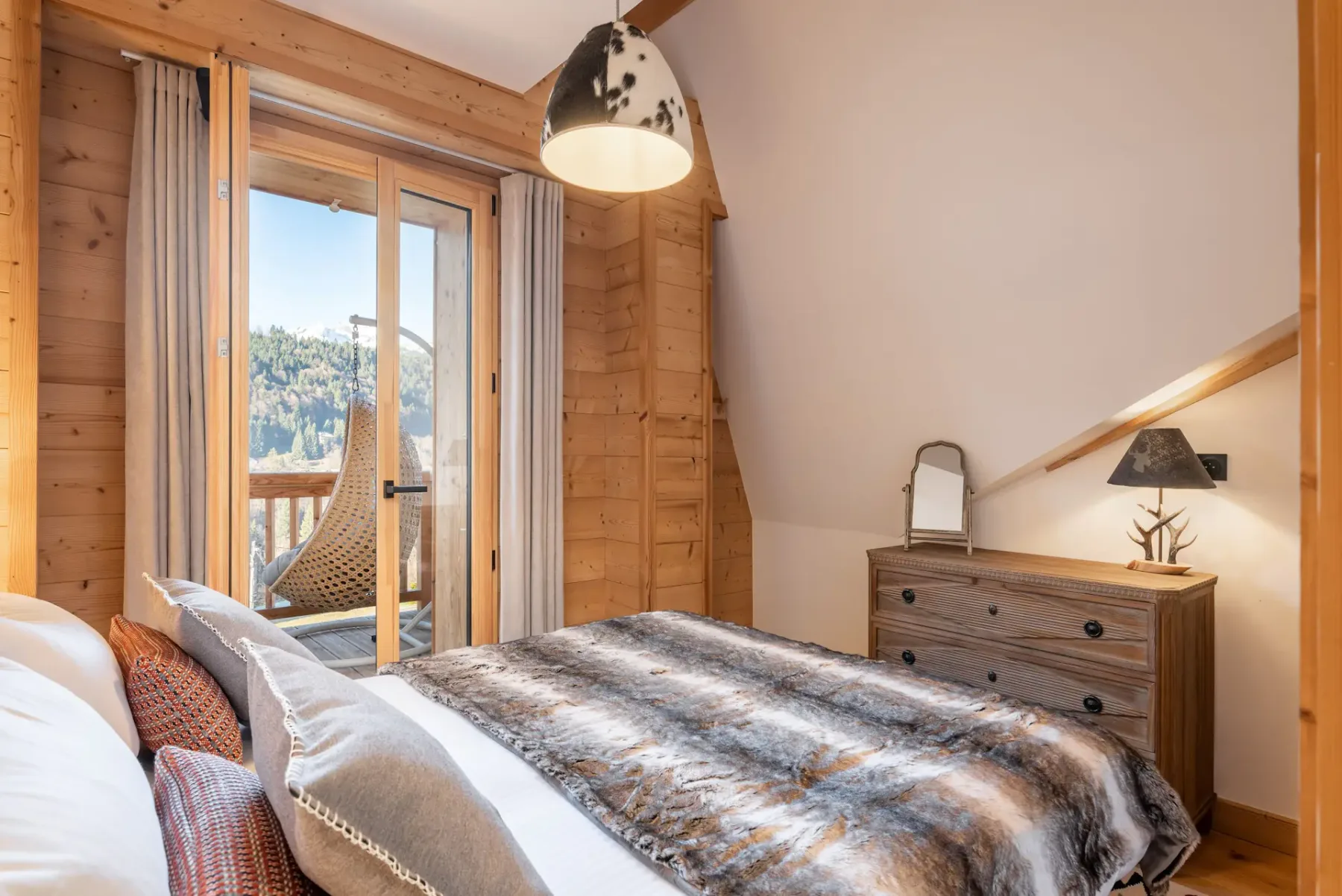 Cozy ensuite bedroom with wooden walls, a bed with fur blanket, a dresser with a lamp and mirror, and a window with mountain view.