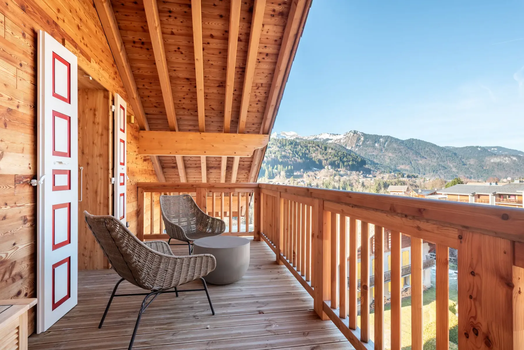 A wooden balcony with two wicker chairs and a round table overlooks a mountain landscape under a clear sky.