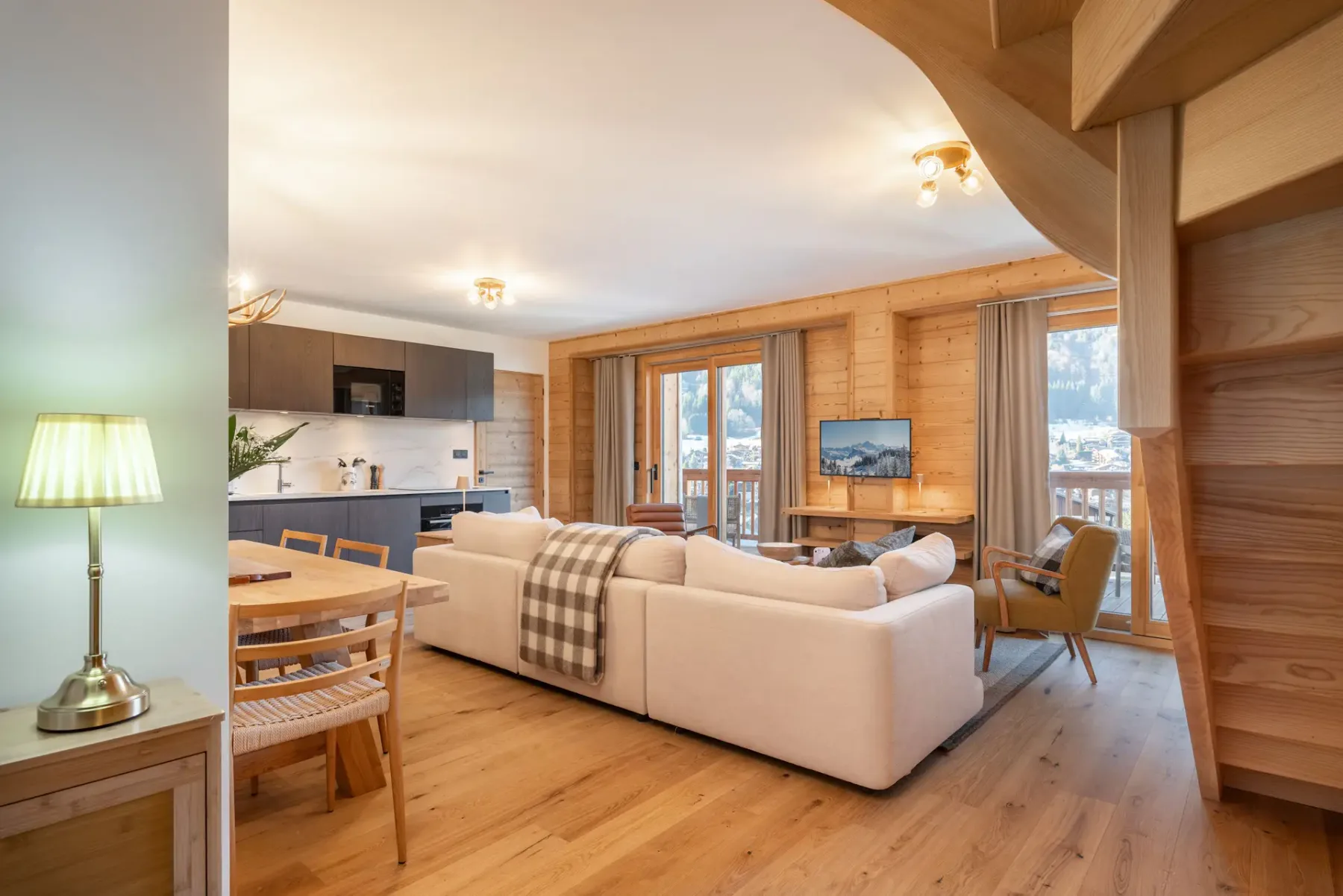 Open-plan living area with a beige sectional sofa, dining table, modern kitchen, wooden flooring and walls, and a staircase, with glass doors leading to a balcony and mountain view.