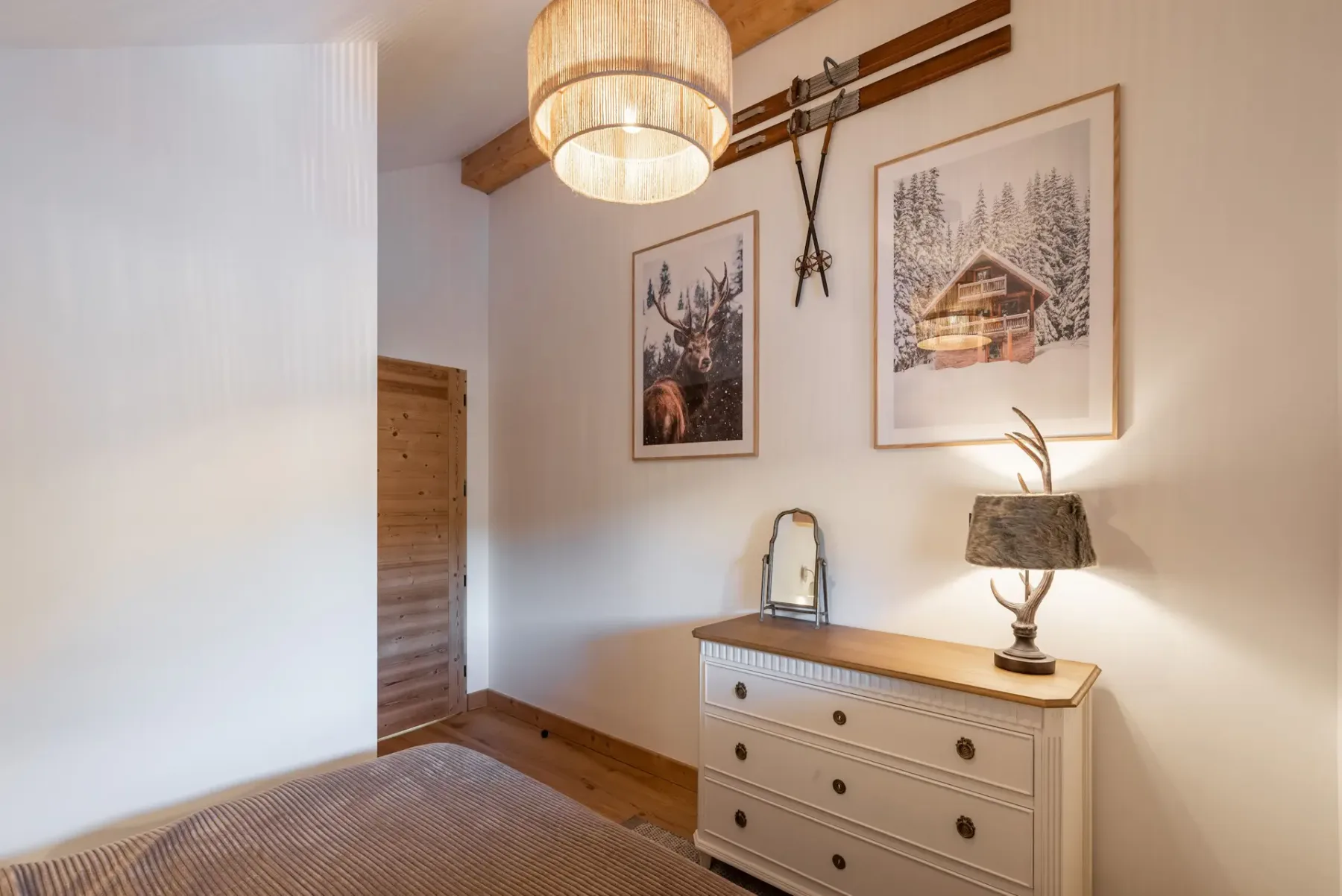 A cozy bedroom with a double bed, plaid pillows, a wooden beam ceiling, a window with a view, and a dresser with a lamp.