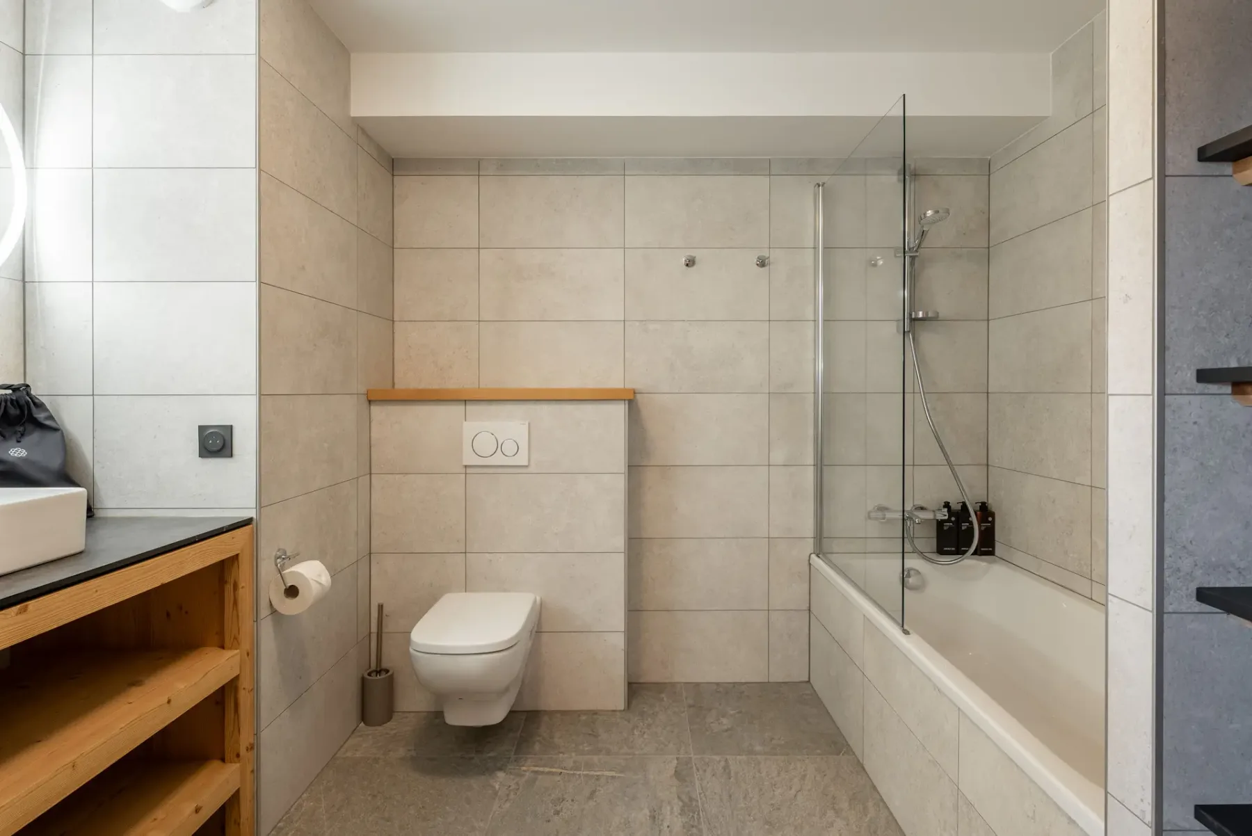 Modern bathroom with gray tile walls, wall-mounted toilet, glass-enclosed bathtub and shower, sink on a wooden vanity, and open shelving.