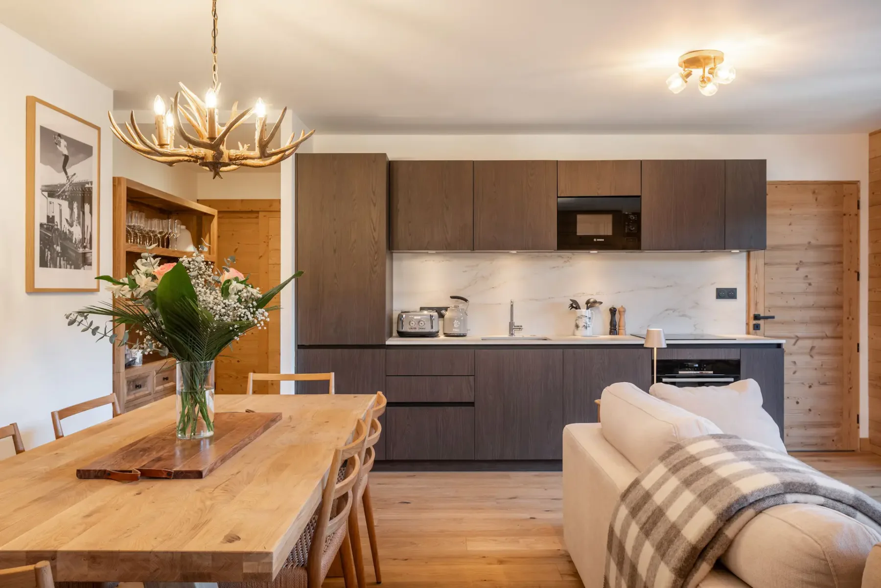 Modern dining area with a wooden table and six chairs, minimalist decor, flower centerpiece, wall art, and open kitchen with dark cabinets in the background.