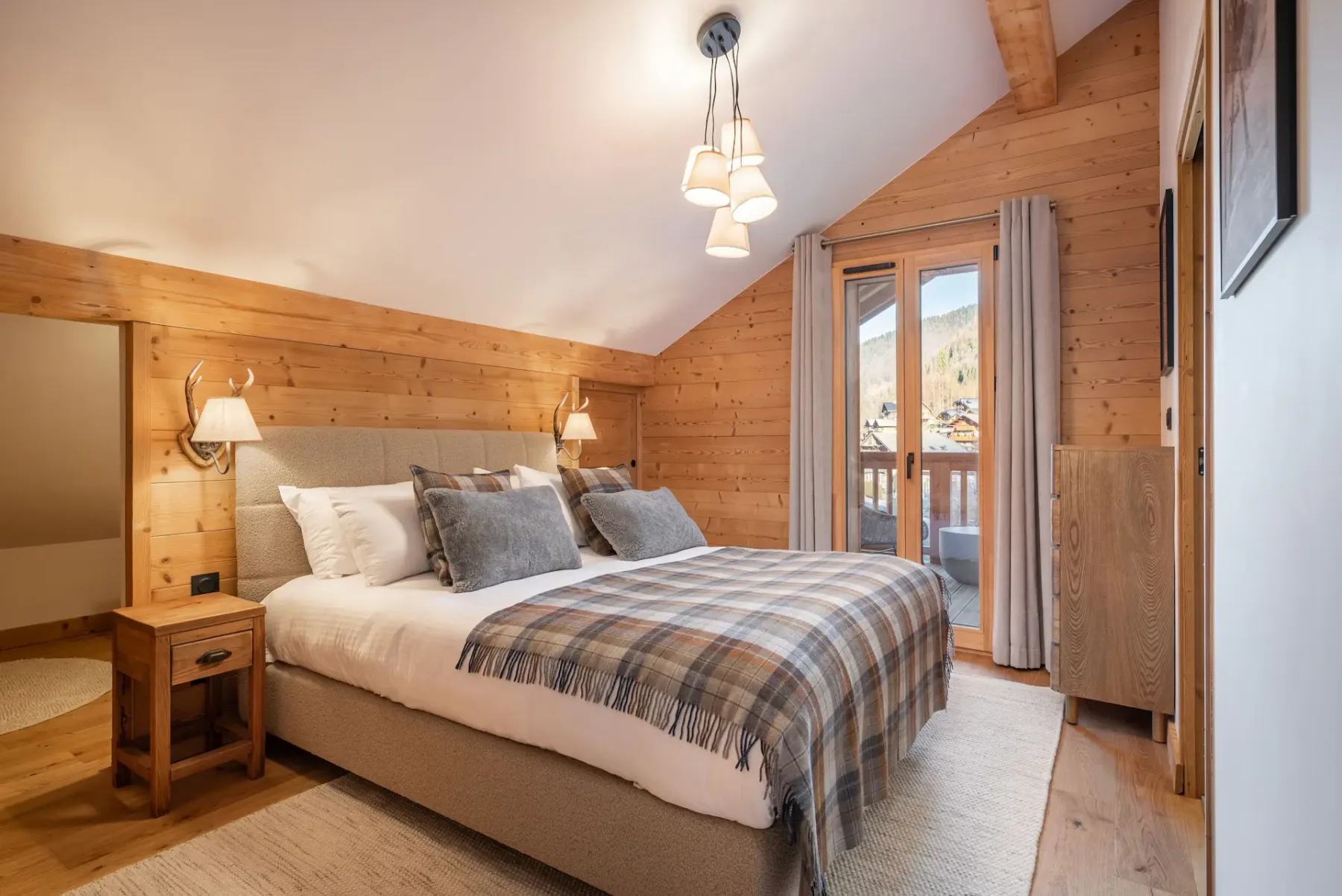 A cozy ensuite bedroom with wood-paneled walls, a double bed with grey and plaid bedding, bedside tables, a mirror, and a balcony door letting in natural light.
