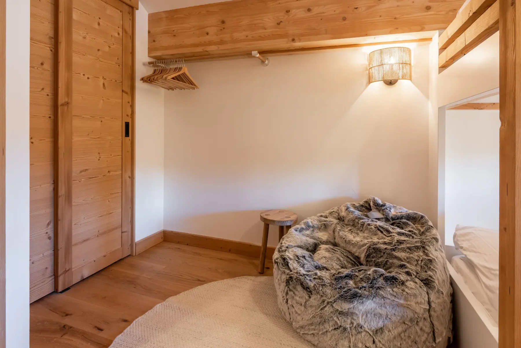 A cozy bedroom with two wooden bunk beds, white bedding, a round furry bean bag chair, a small wooden stool, and warm ambient lighting.